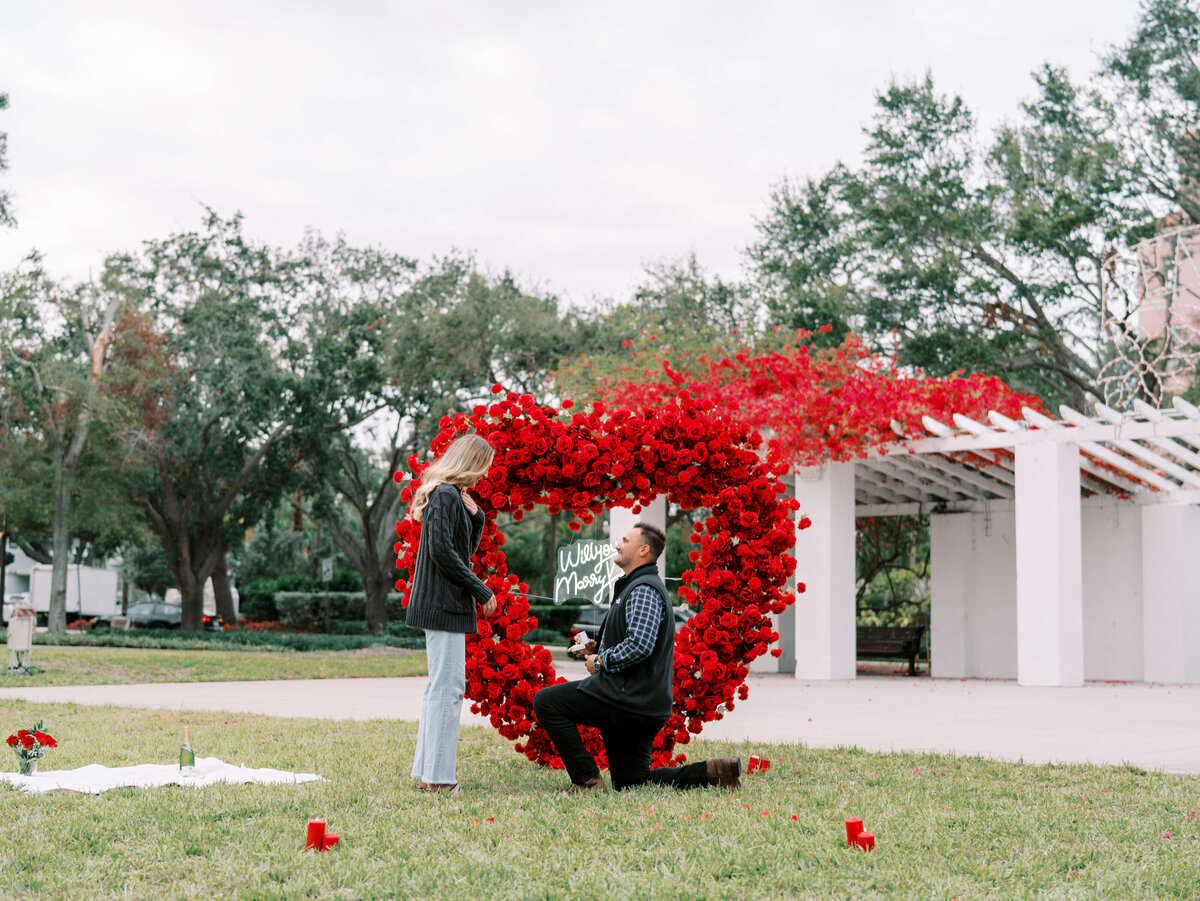 Man proposing to a woman in front of a red floral arch at North Straub park