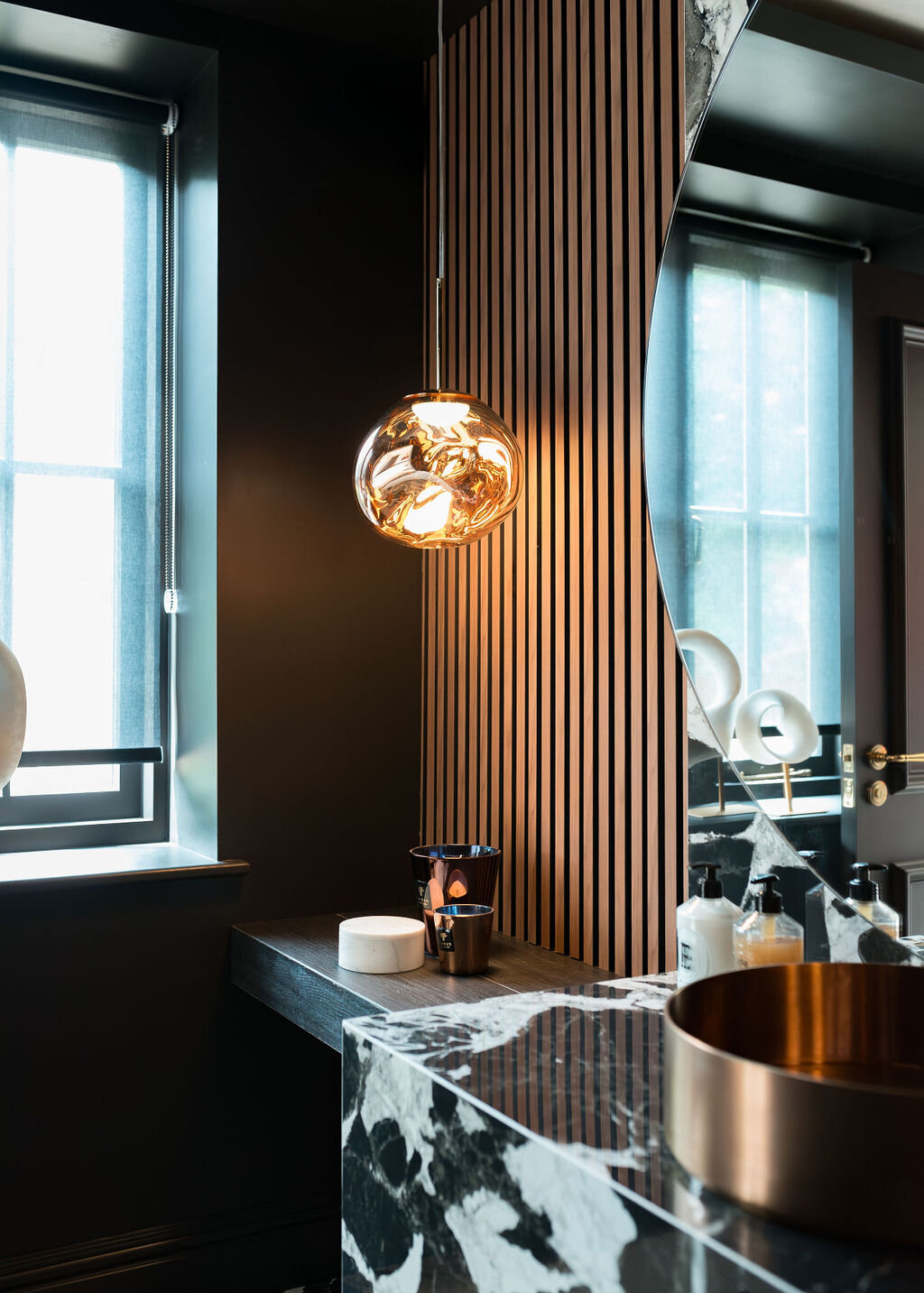 A modern bathroom with a large round mirror, black and white marble sink and countertop, wooden slatted wall, and pendant light fixture. To the left, light flows in from a thinly shaded window.