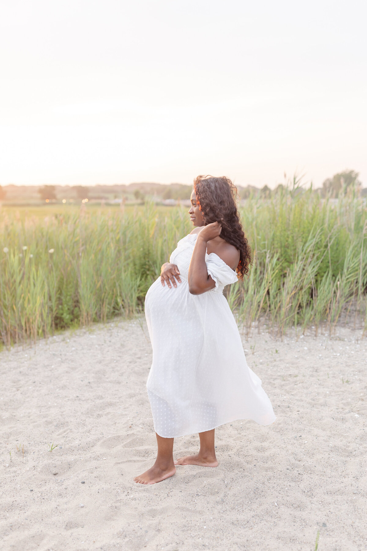 a beautiful african american mother stands on beach dunes with tall grasses in the background, while wearing a white dress, ready for her new baby to come into the world, captured by Connecticut Maternity Photographer Oak and Lore Studios