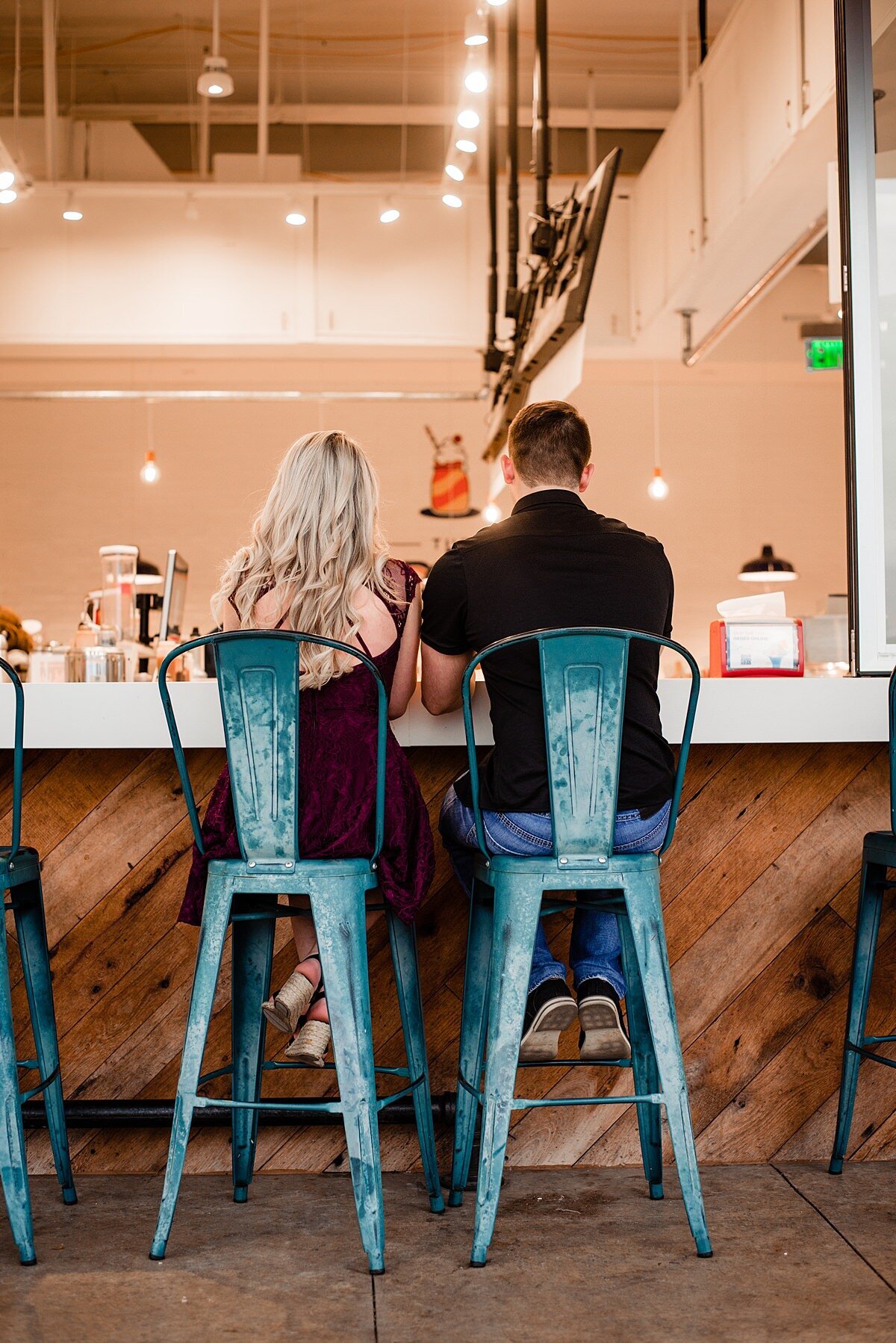 The bride and groom sit in tall teal blue chairs at a soda fountain sharing a drink. The bride is wearing a maroon dress and the groom is wearing dark wash jeans and a black shirt.