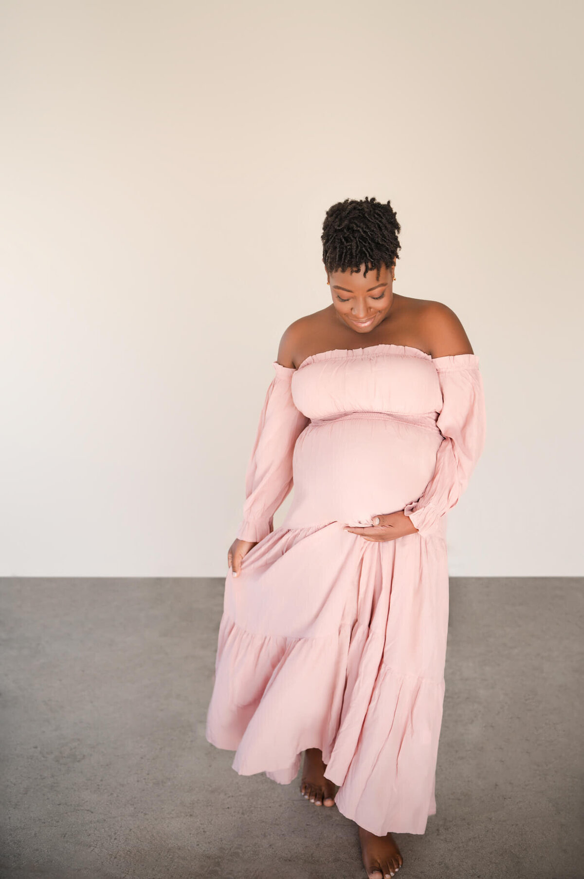 pregnant woman in pink dress holding belly while walking in a studio smiling with short hair