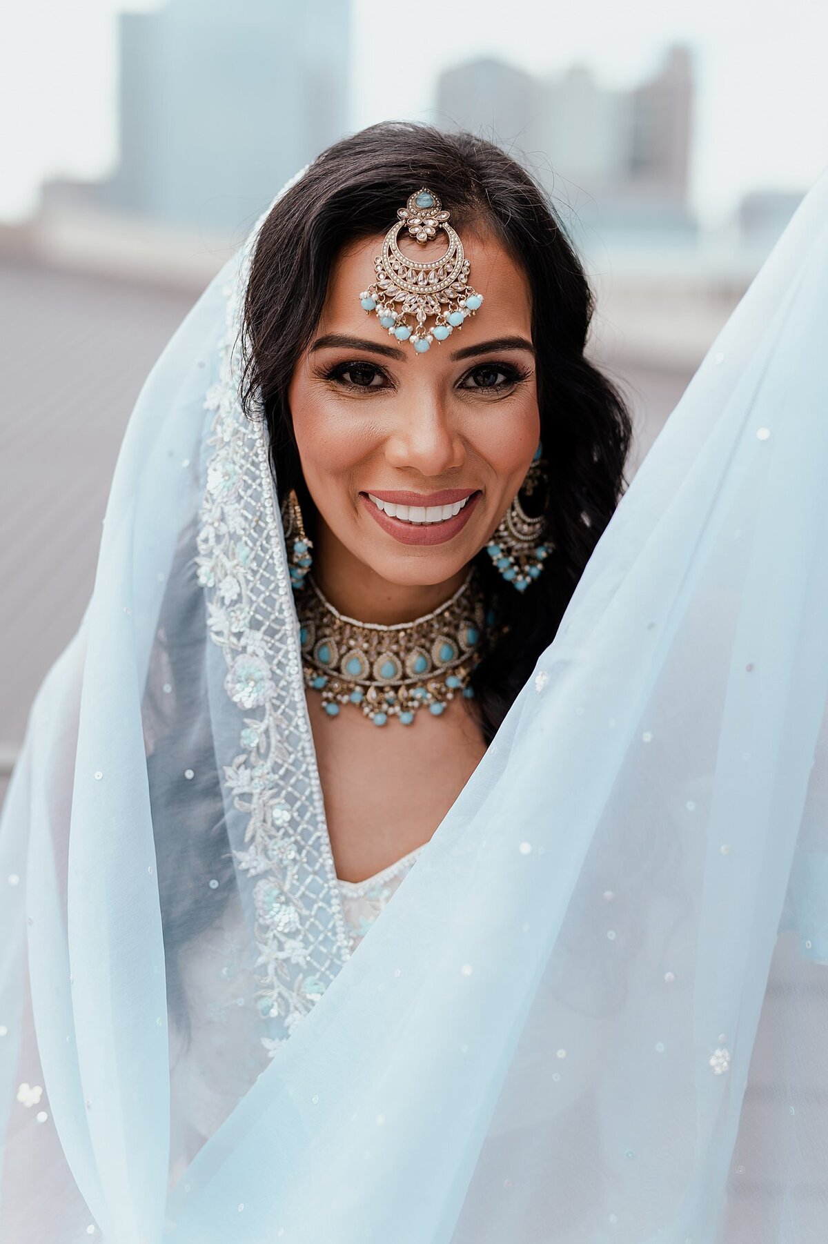 The Indian bride holds up her light blue dupatta as she smiles at the camera displaying her light blue and gold wedding jewelry.