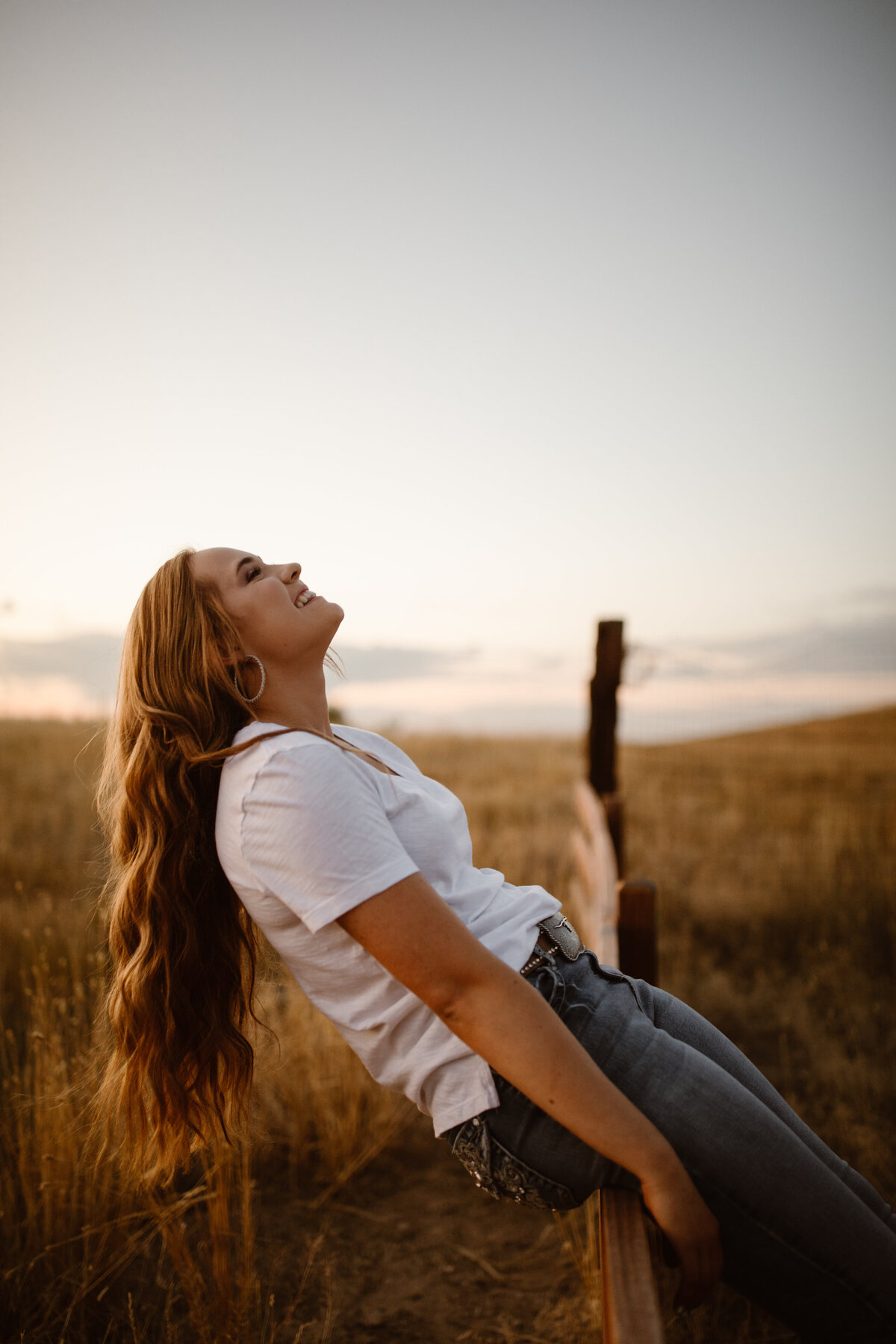 Senior girl with long wavy hair leans back on a wooden fence, smiling up at the sky during golden hour in a dry Colorado field.