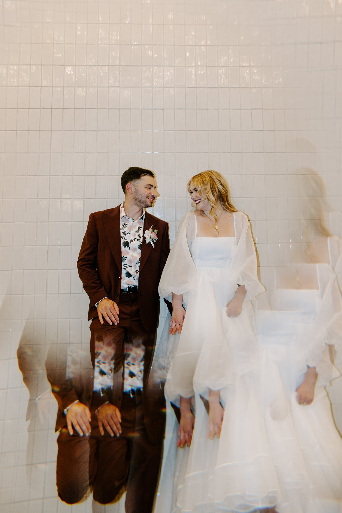 A couple holds hands in front of a white tile wall.