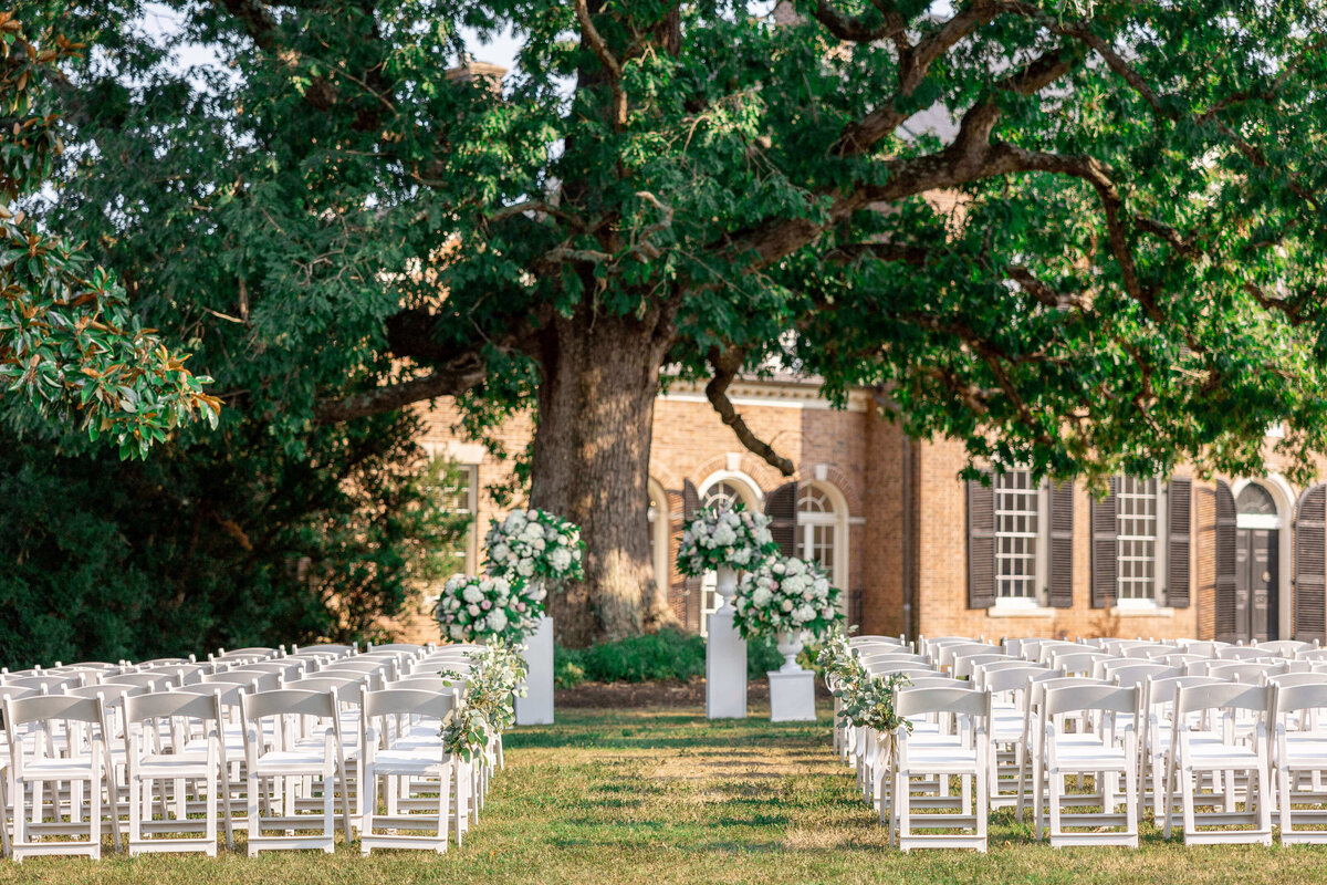 White chairs are arranged in rows facing a large tree, with a historic brick building in the background. Elegant floral arrangements with white flowers adorn the aisle, creating a picturesque outdoor wedding setting.