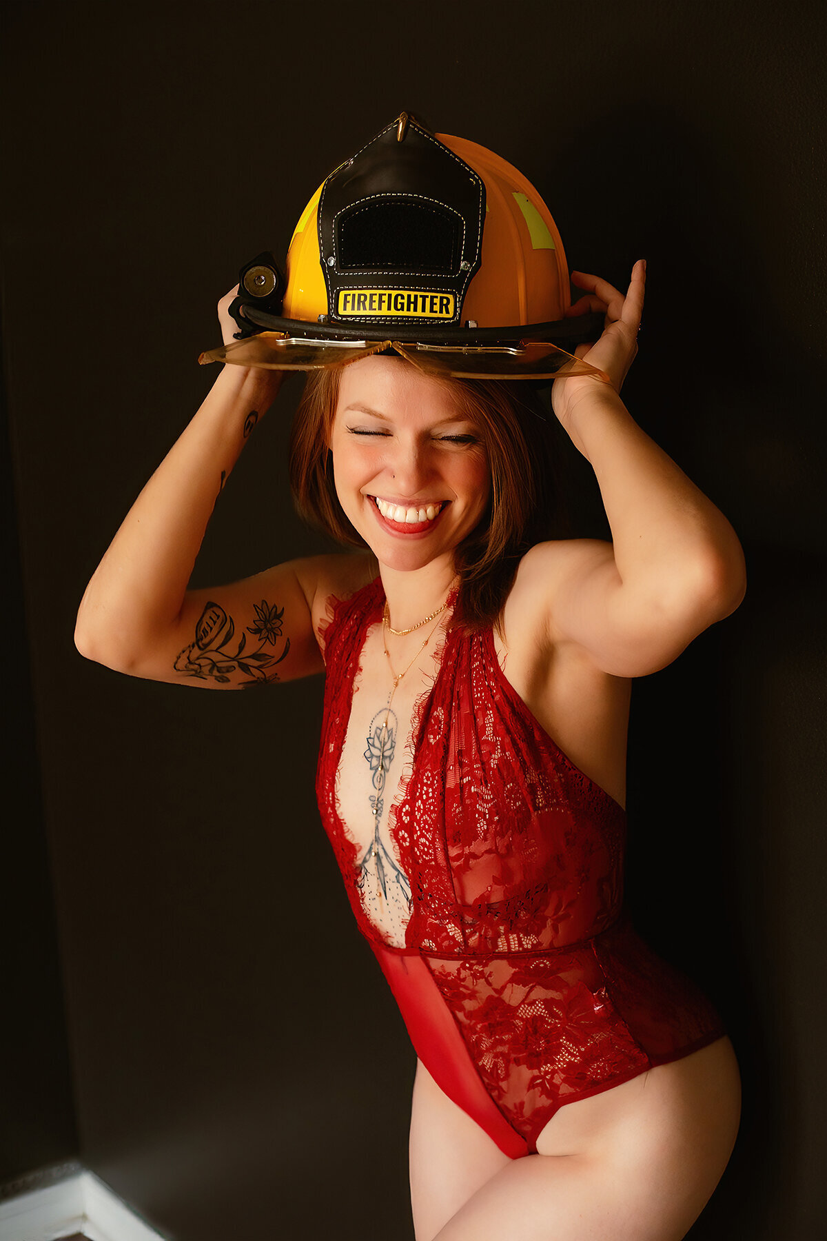 A woman wearing red lingerie smiles and laughs while wearing a fireman's hat during a boudoir photoshoot.