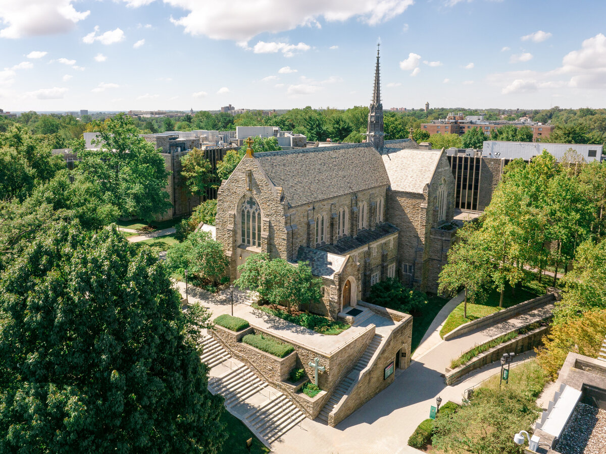 Aerial view of a historic stone chapel with large arched windows and a tall spire, surrounded by lush green trees and pathways. The sky is clear with a few fluffy clouds.