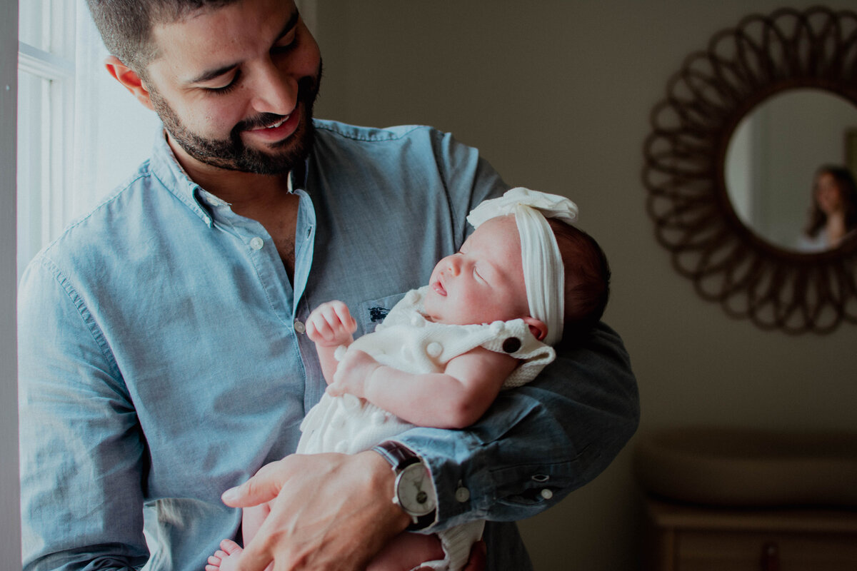 Dad and baby girl standing by a window during ad lifestyle newborn photoshoot in CHicago, IL