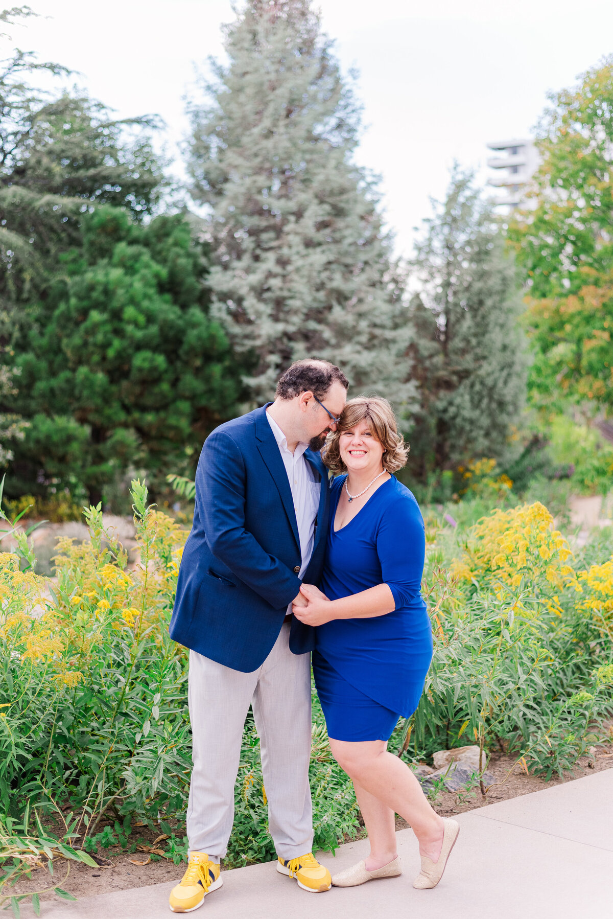 A man in a blue jacket and a woman in a blue dress stand side by side as the man nuzzles her ear and she looks at the camera while they both hold hands, captured by Denver wedding photographer