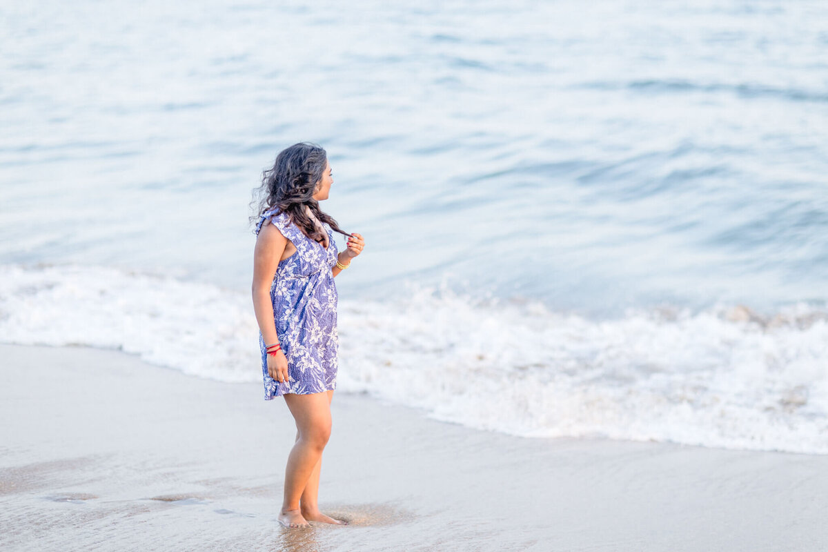 A person with long hair stands on a sandy beach, wearing a blue and white dress. They are looking at the sea, with gentle waves lapping at their feet. The ocean stretches into the distance under a clear sky.