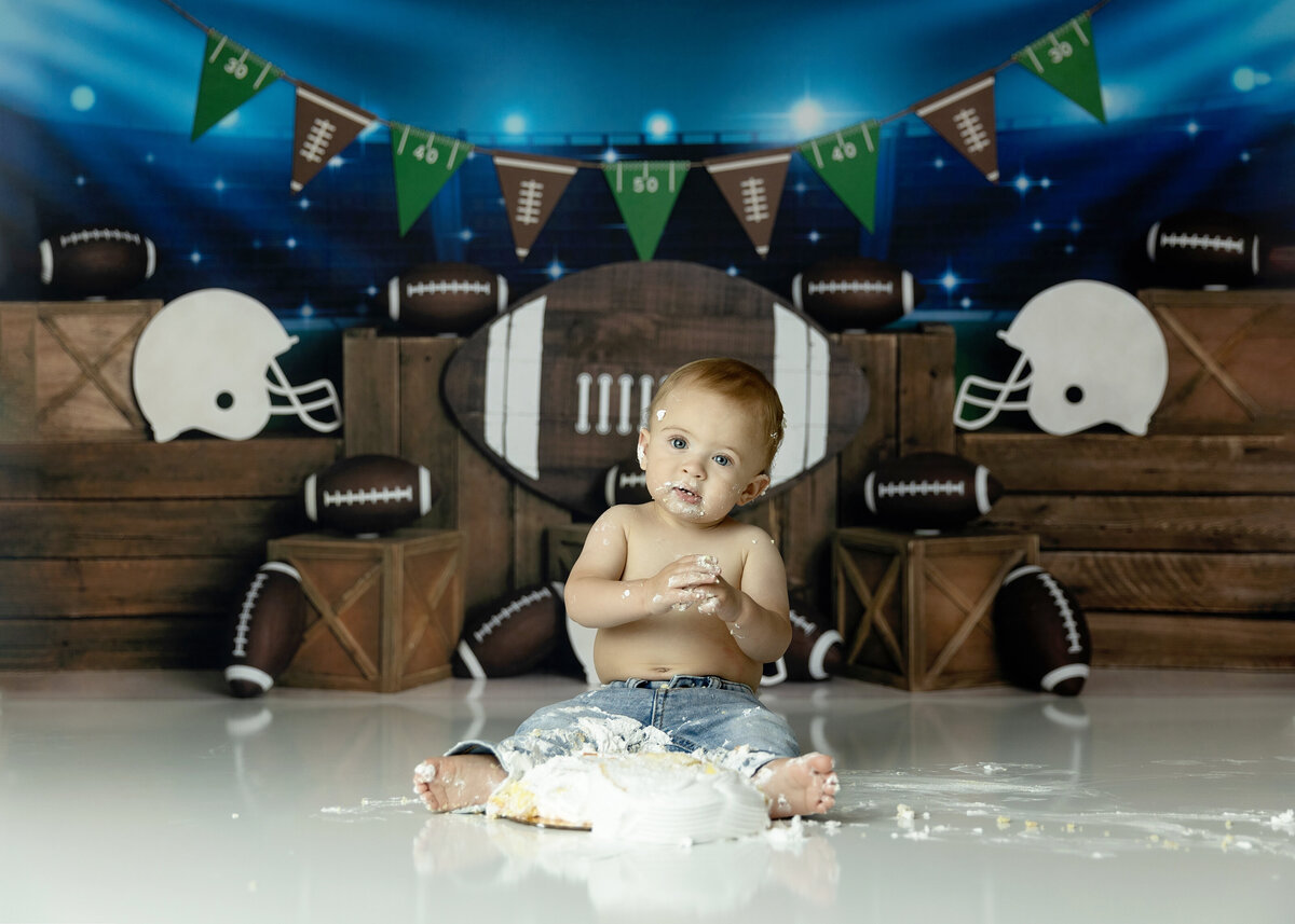 A toddler boy in jeans sits in a football themed studio smashing a cake