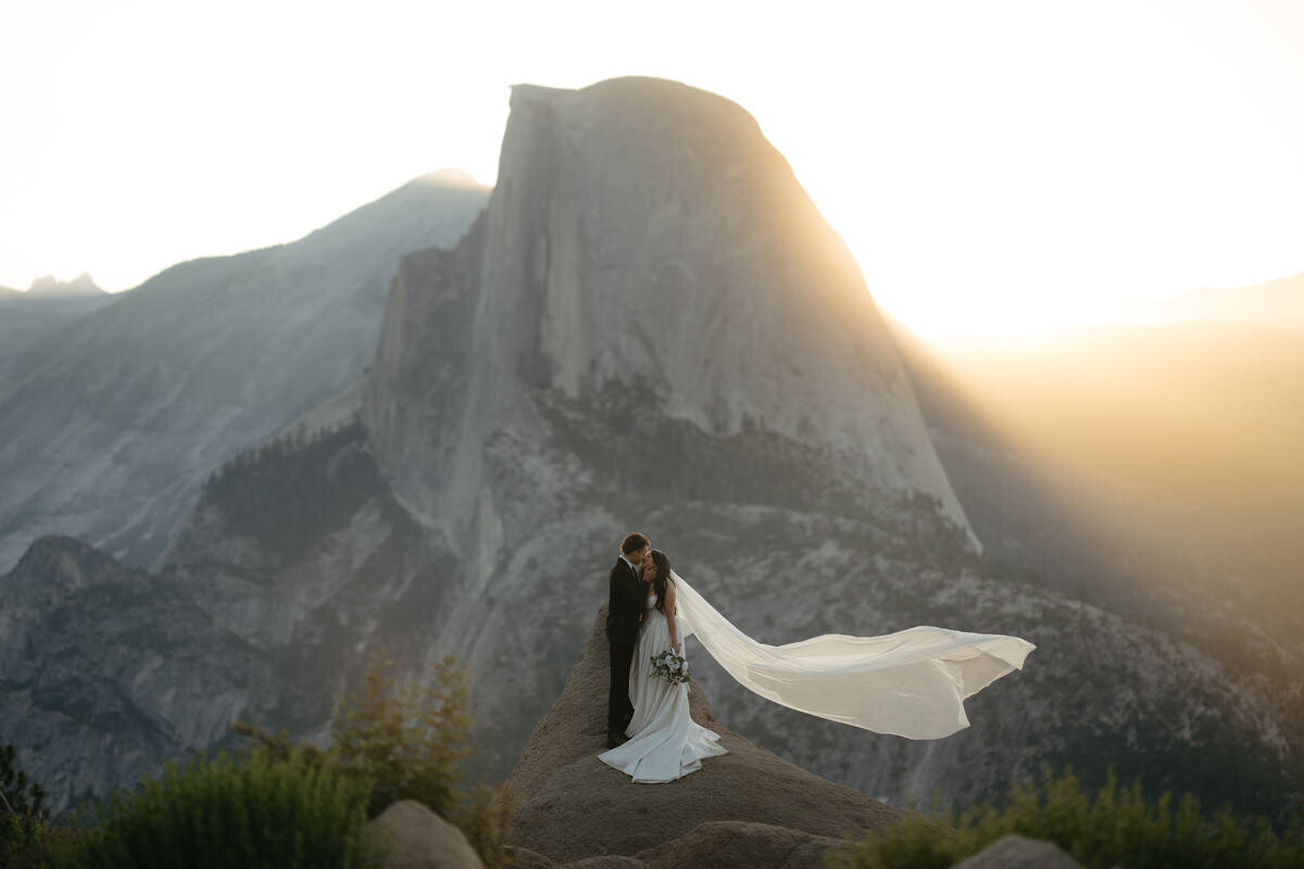 Beautiful sunrise Glacier Point Wedding, bride's veil is flowing the wind illuminated by the sun rays from behind Half Dome