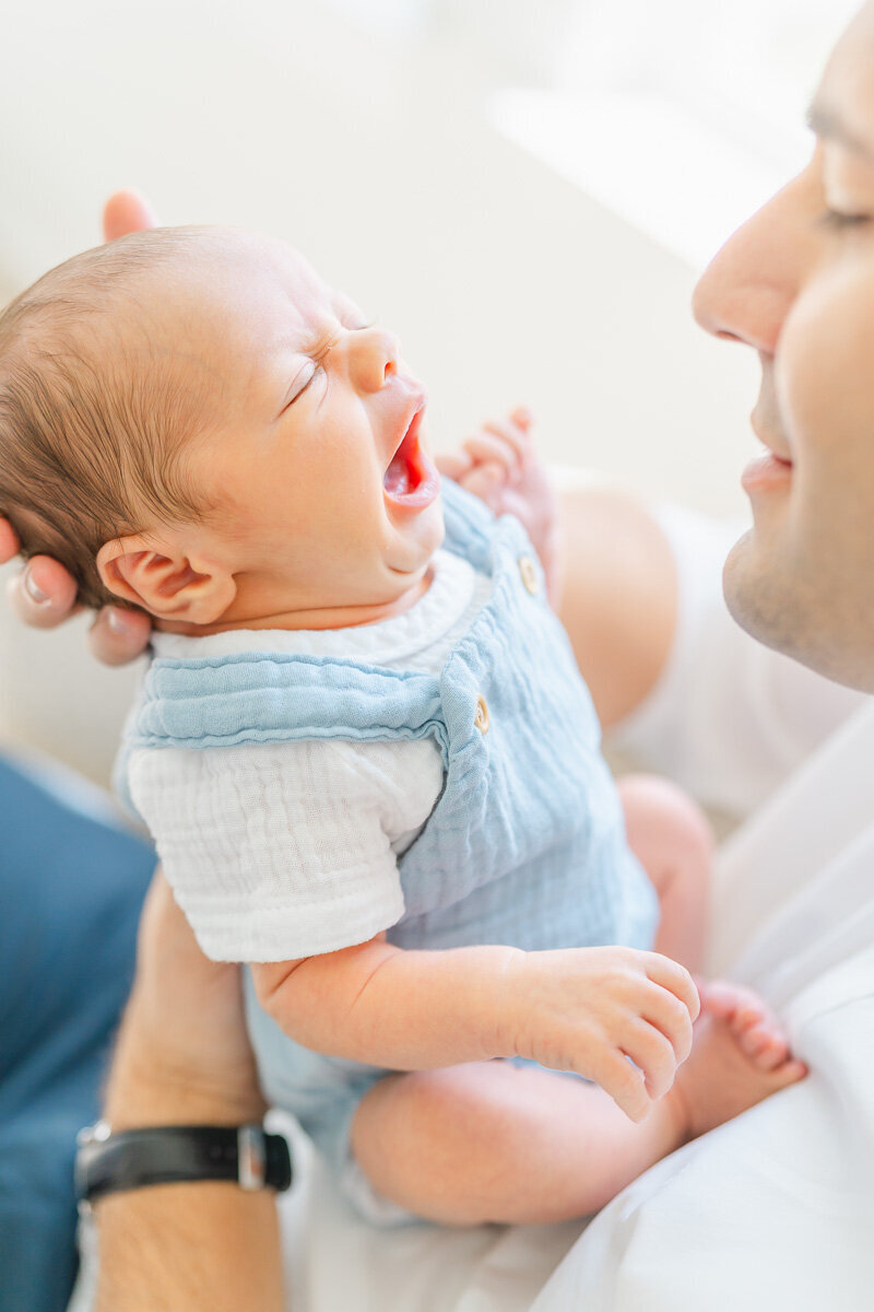 a newborn dressed in a blue romper is held by his father and yawns while captured by a Leander photographer.
