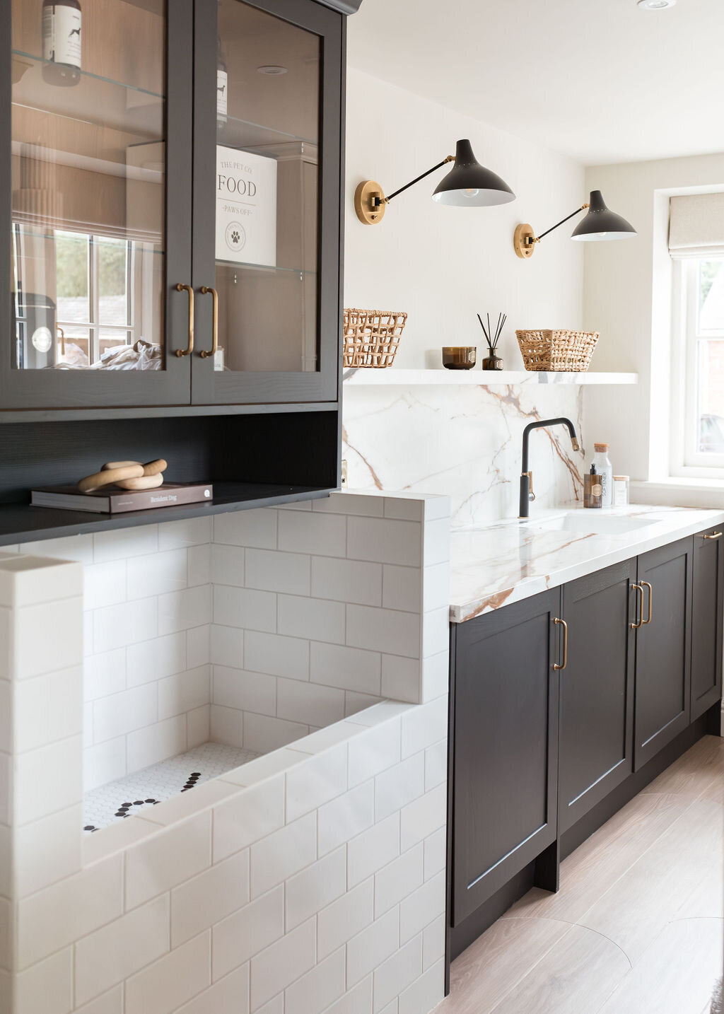 A modern dog wash room with black cabinets, a large white subway tiled washing station and white marble countertops. A black built-in shelf sits above the washing station. A shelf with wicker baskets sits above the counter, along with two black lights.