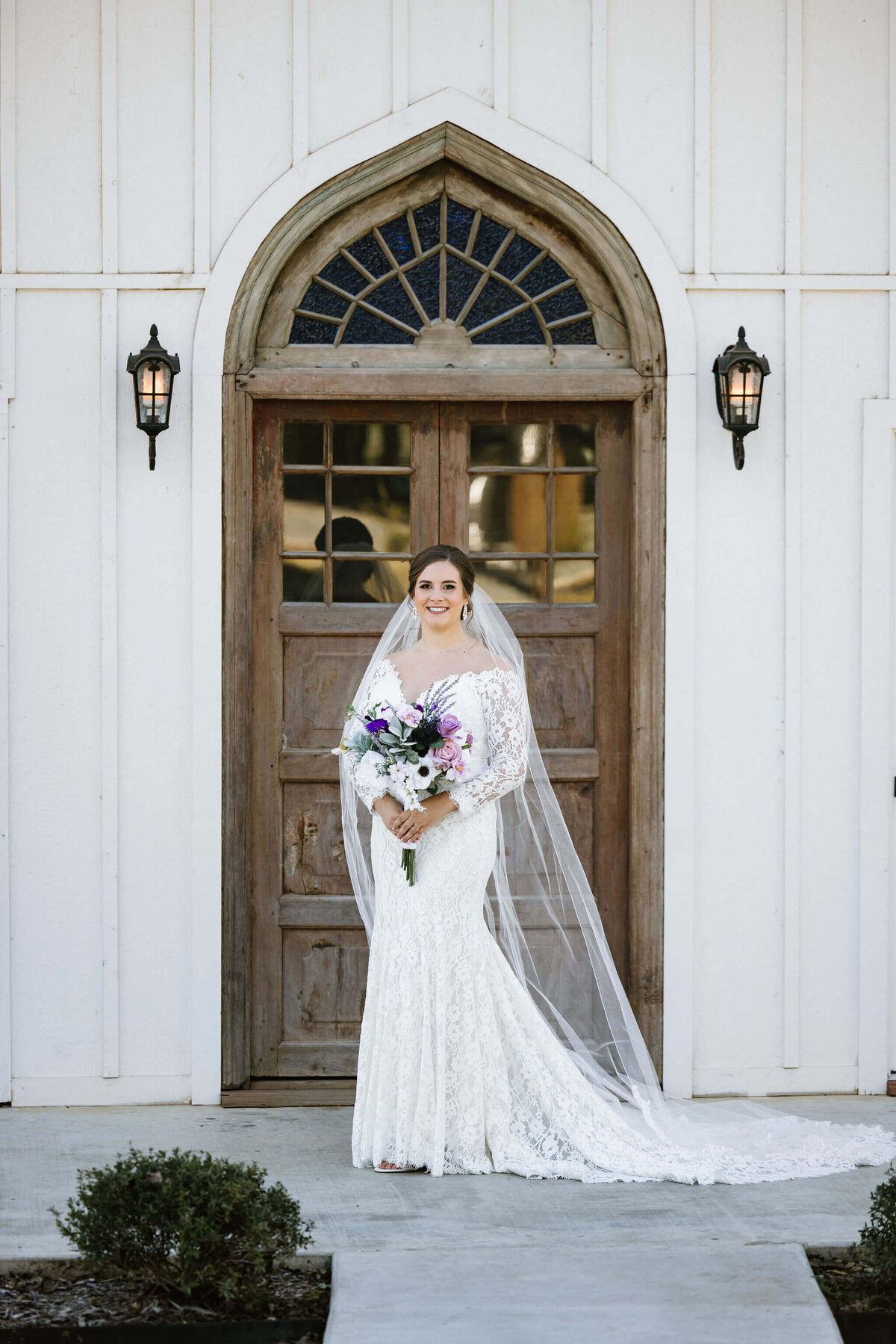 bridal portrait of bride in vintage lace white wedding gown and cathedral veil holding spring colored bridal bouquet while standing in doorway of Four Fifteen Estates wedding venue in New Boston, TX