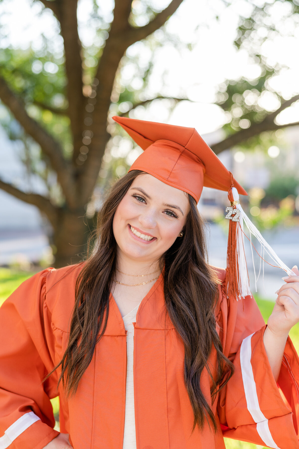 Caldwell High School senior girl in orange gown and hat while holding tassel and smiling in Downtown Brenham