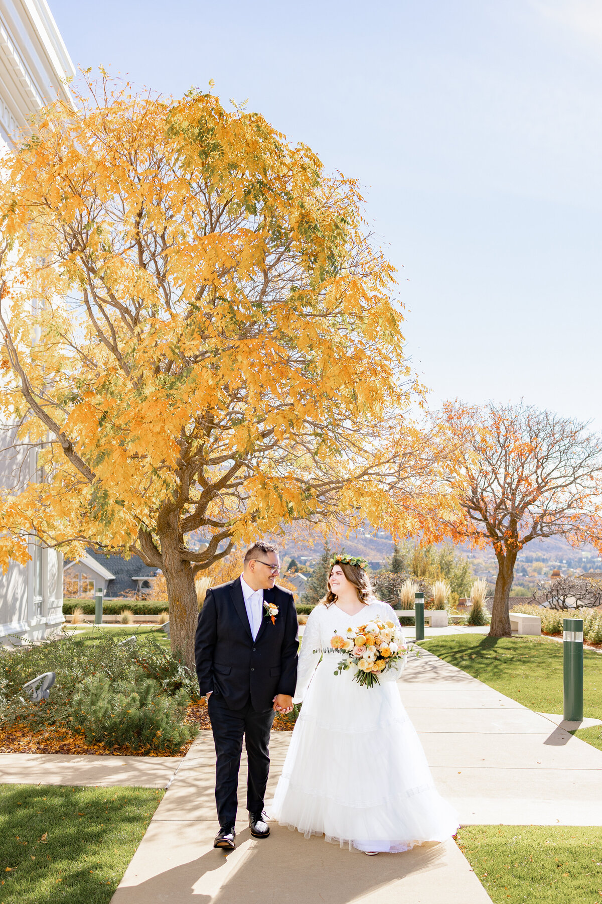 A gorgeous bride and her new husband walk hand in hand down a pathway lined with orange Autumn trees