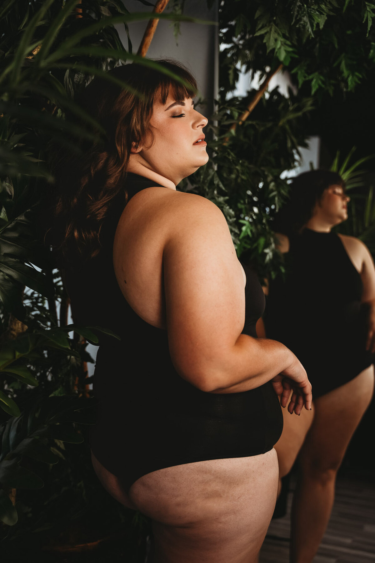 beautiful plus size brunette woman wearing a black body suit, standing beside greenery plants and in front of a mirror