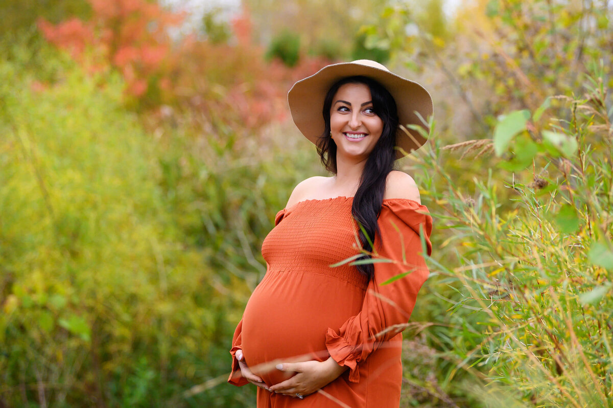 Prenant momma-to-be wearing an orange maternity dress and beige hat for her outdoor fall maternity photography session in Burlington, Ontario