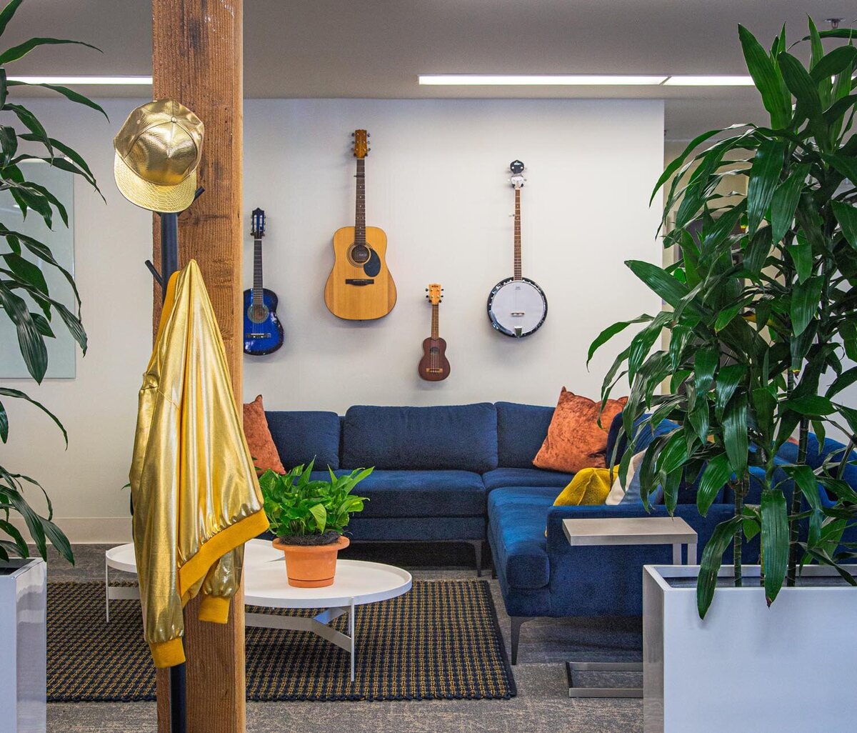 Lounge with blue sectional sofa, wall-mounted guitars, banjo, and ukuleles, surrounded by large indoor plants.