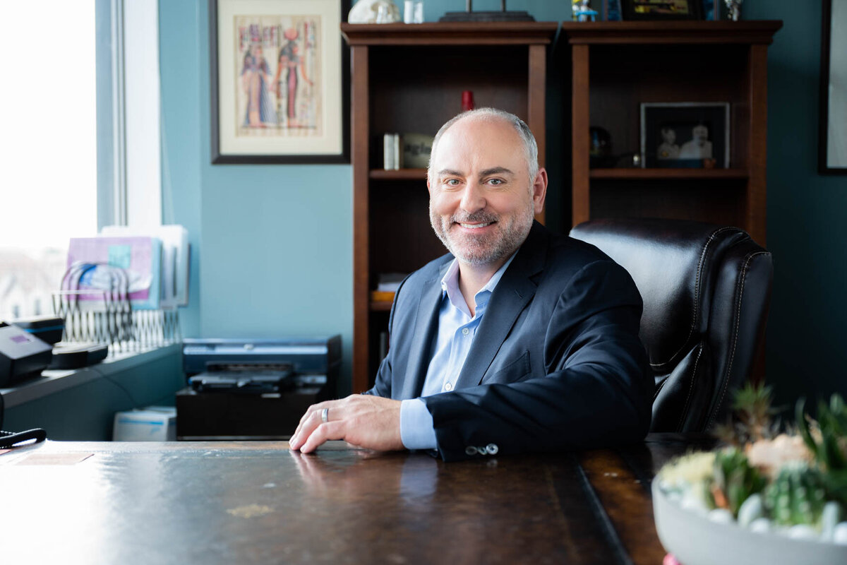 A professional headshot featuring a person seated at a desk in a bright office space. The individual wears a navy suit jacket over a light blue shirt, paired with a friendly smile and confident posture