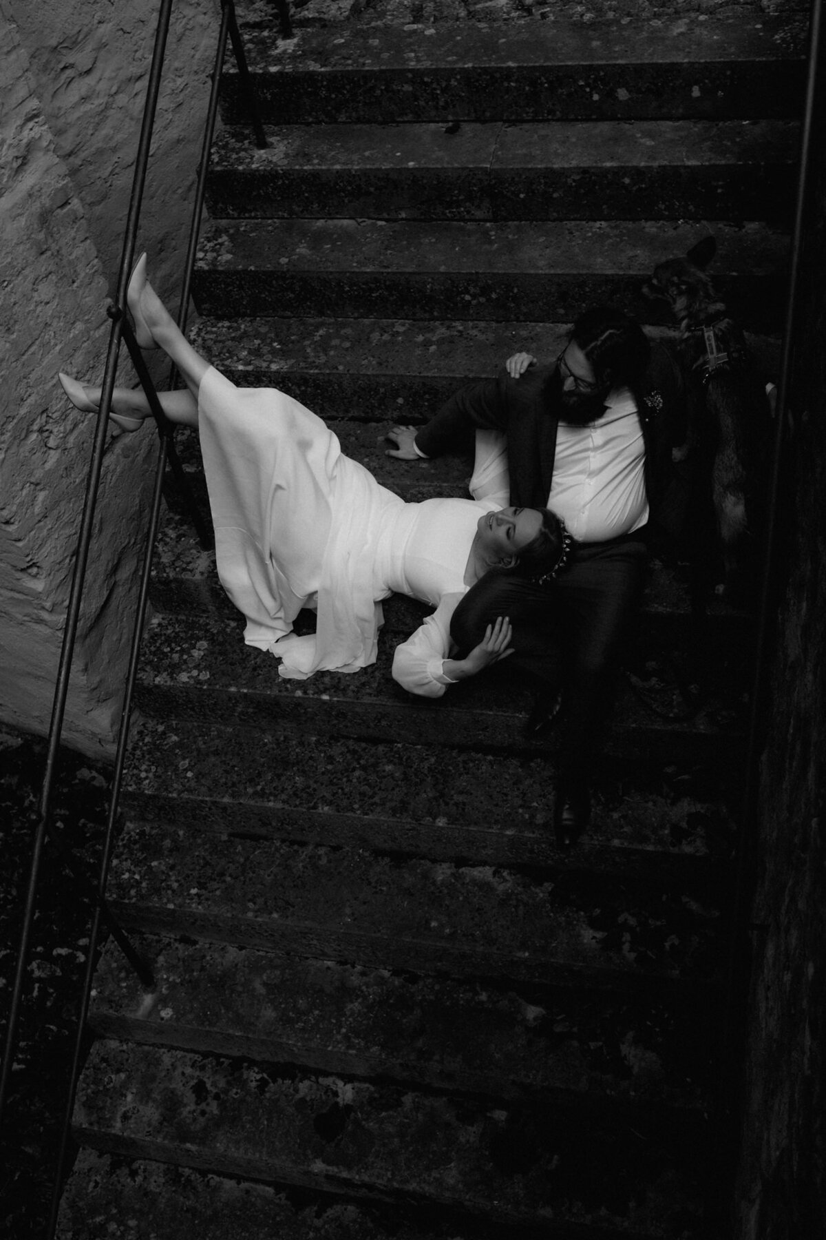 Auf einer Steintreppe liegt die Braut mit leicht angehobenen Beinen auf ihrem sitzenden Mann.