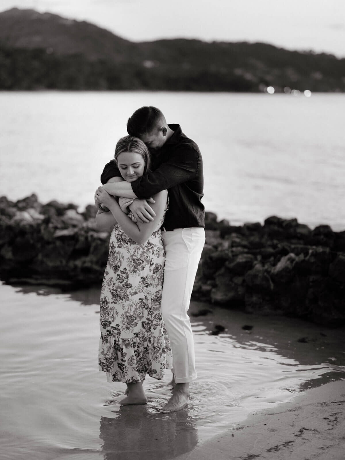 Black and white photo of the engaged couple standing on the seashore, with the man hugging the woman from behind
