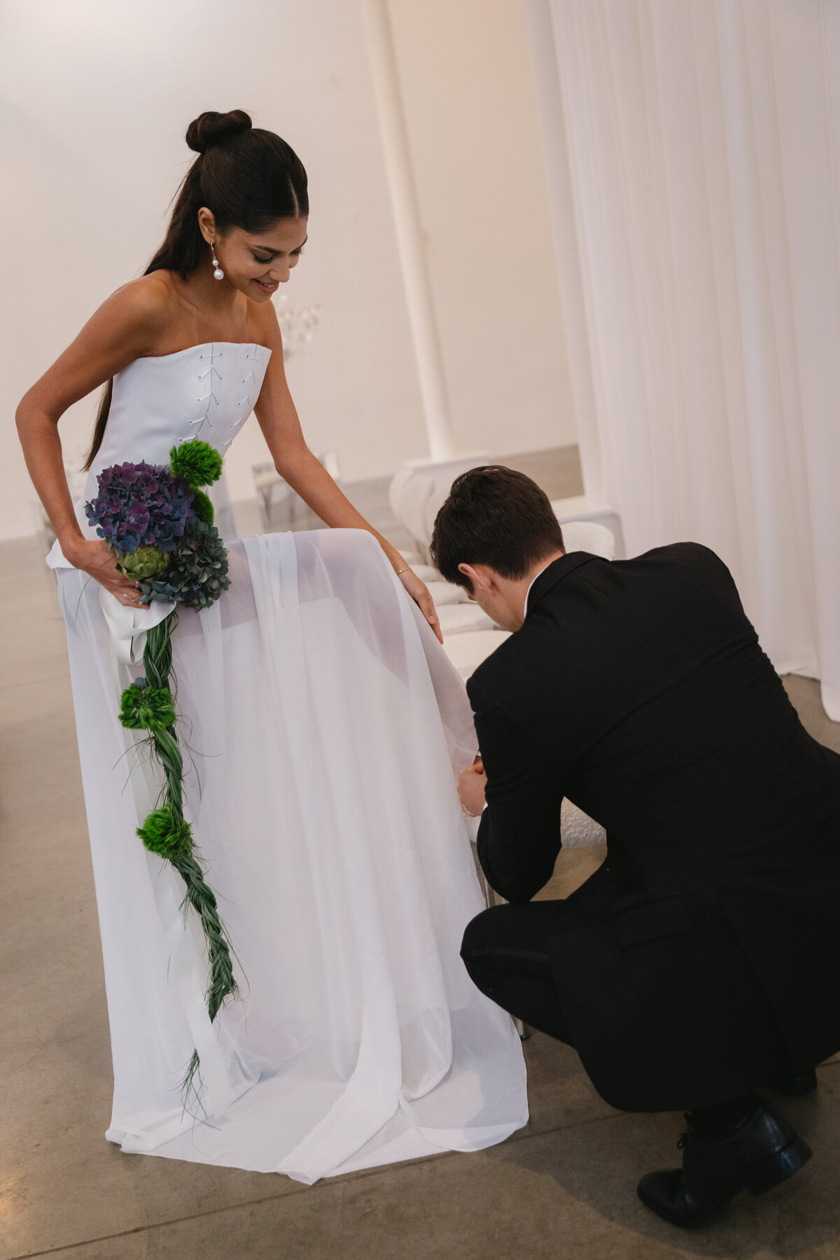 Groom helping adjust the bride’s flowing dress during a candid, natural moment in a minimalist wedding setting.