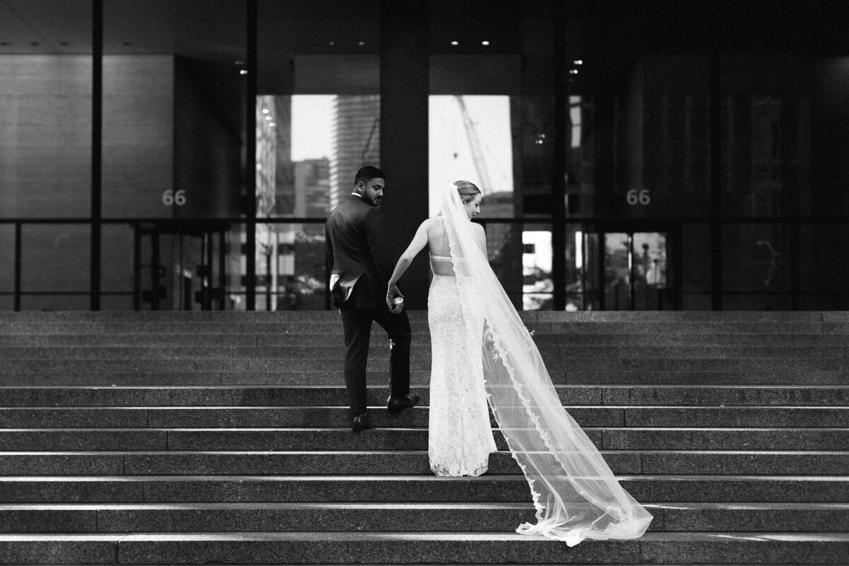 Bride and groom holding hands, walking up steps outside a building at Steam Whistle Brewing. The bride wears a long veil. Captured by Jennifer van Son, a talented wedding photographer in Toronto, this black and white image beautifully preserves their cherished moment.