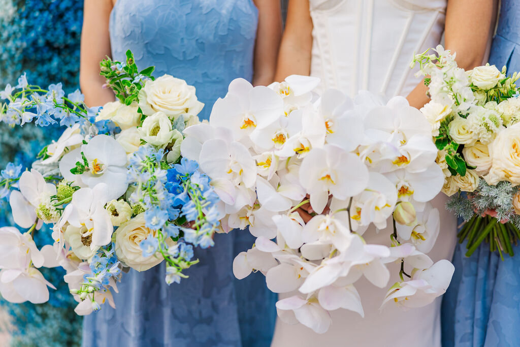 A stunning bridal party with bride dressed in elegant blue and white, each bridesmaid holding lush bouquets of fresh orchids and roses. The combination of soft florals and sophisticated color tones creates a timeless and refined wedding aesthetic, perfect for couples seeking a classic yet modern celebration.