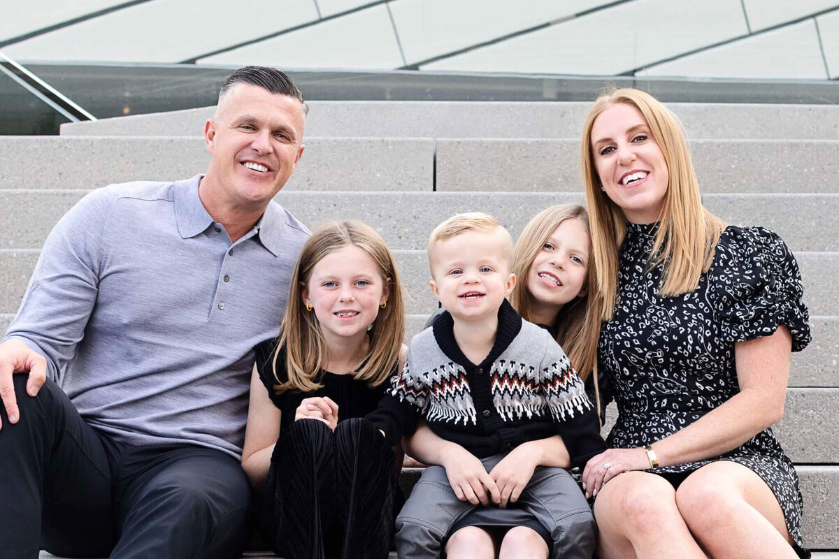 A mom, dad, and three young children wearing gray and black smile at the camera while sitting on grey steps.