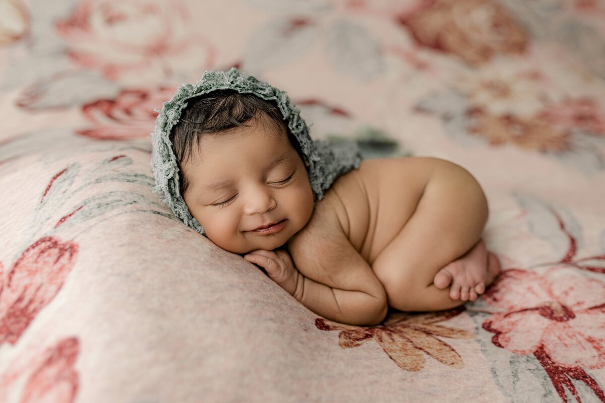 studio newborn session with a flower backdrop and a newborn baby girl wearing a green bonnet and curled up in a cute little pose