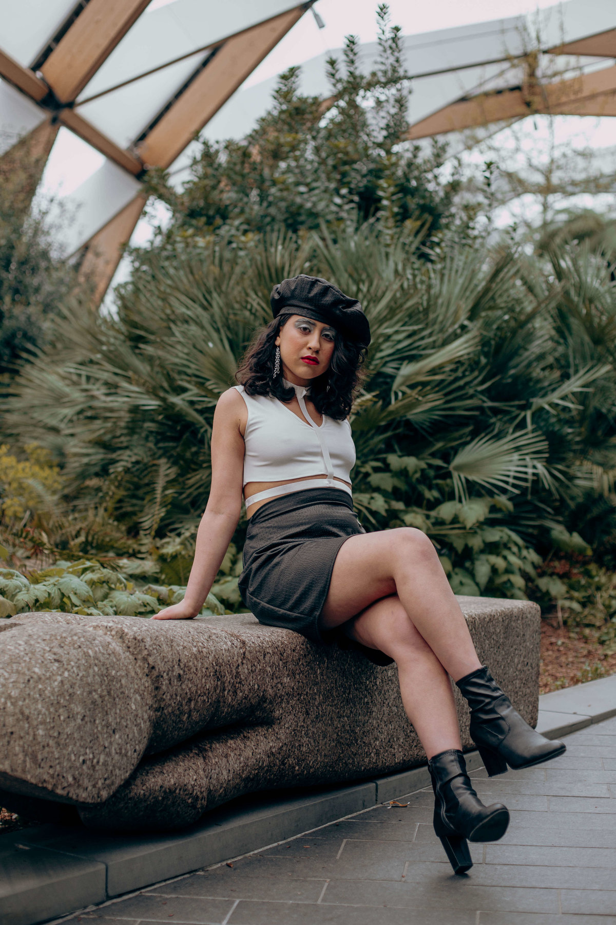 Cherie wearing a black and white leather outfit with red lipstick, sat on a stone bench infront of a green bush.