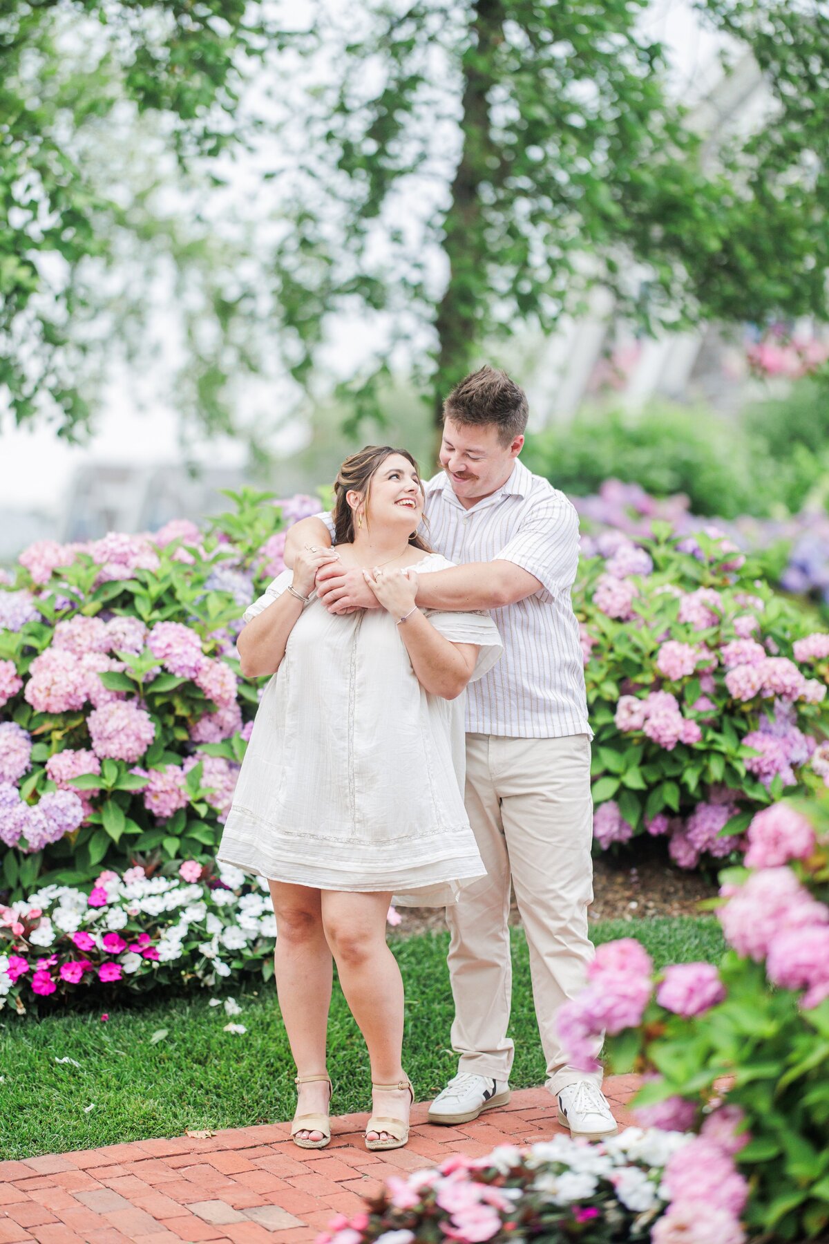 Man standing behind a woman with his arms wrapped around her shoulders, both smiling, surrounded by colorful hydrangeas and white flowers at Chatham Bars Inn in Chatham, Massachusetts — representing the portfolio of a Cape Cod engagement photographer.
