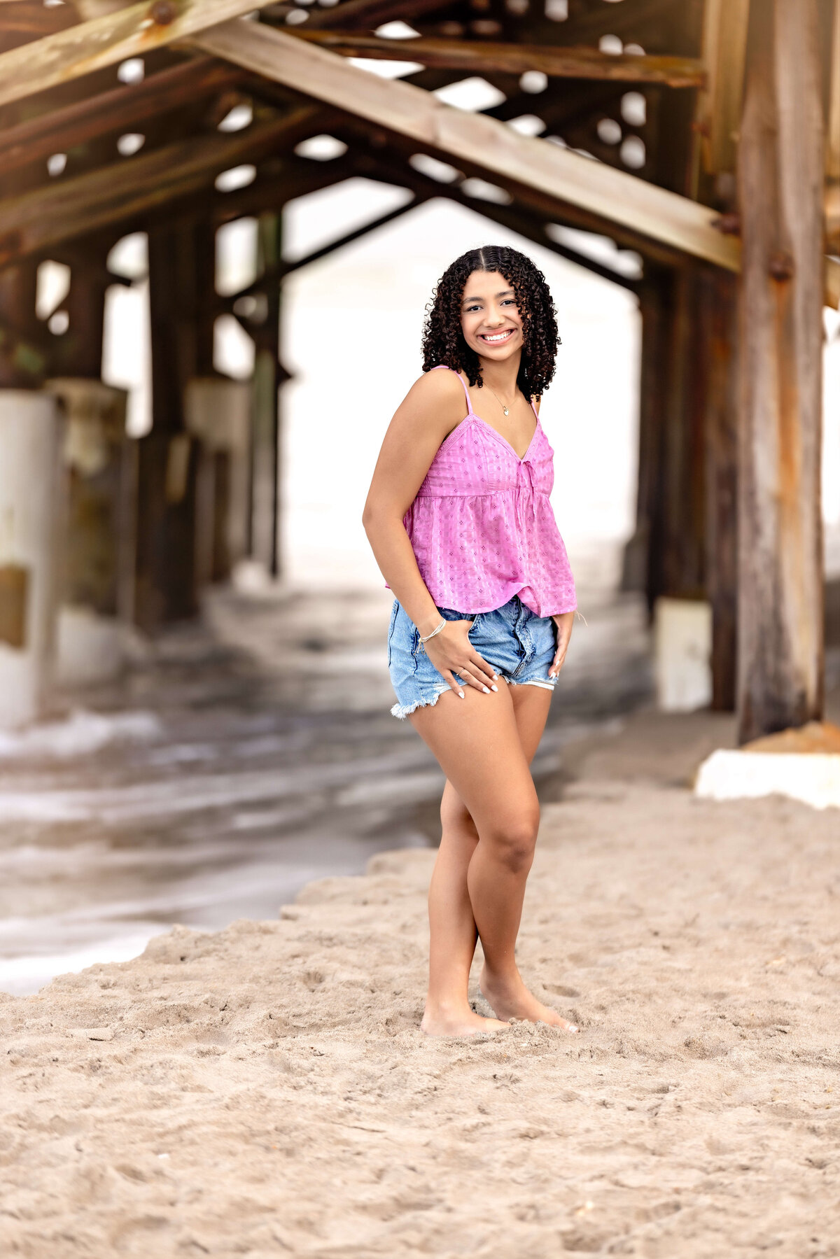 Windermere senior session with a girl standing under Cocoa Beach Pier, pink top and jean shorts, smiling with soft light and ocean behind her.