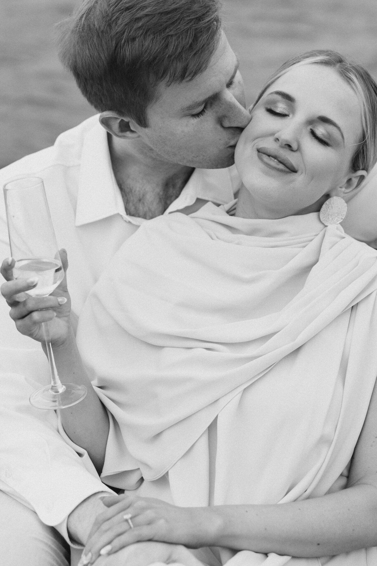 Guy kisses woman on cheek while she holds her champagne glass on their boat engagement session on Lake Oconee