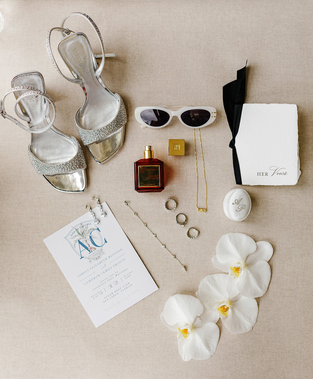 A flatlay of wedding details including silver heels, an invitation suite, orchids, and a perfume bottle, showcasing the bride's style and sophistication, captured by Claudia Amalia Photography.