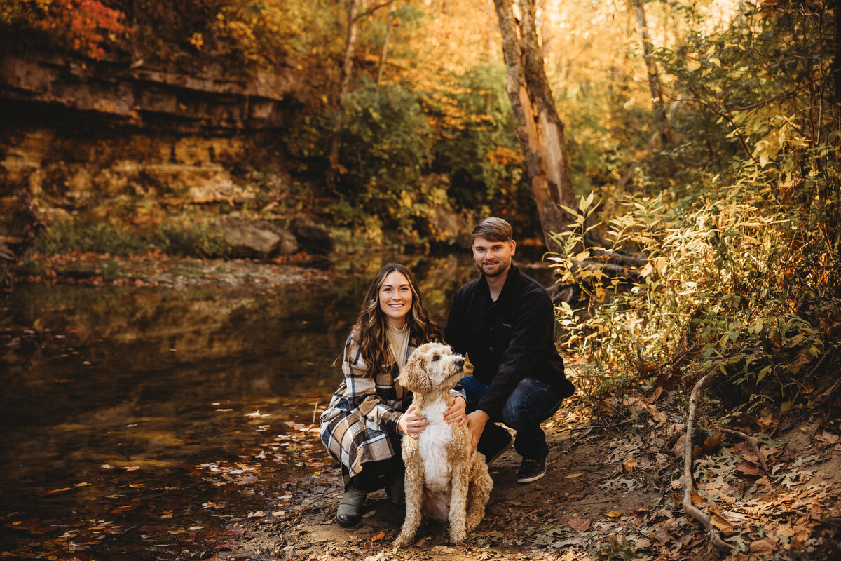 A lovely couple crouches behind there dog by a creek