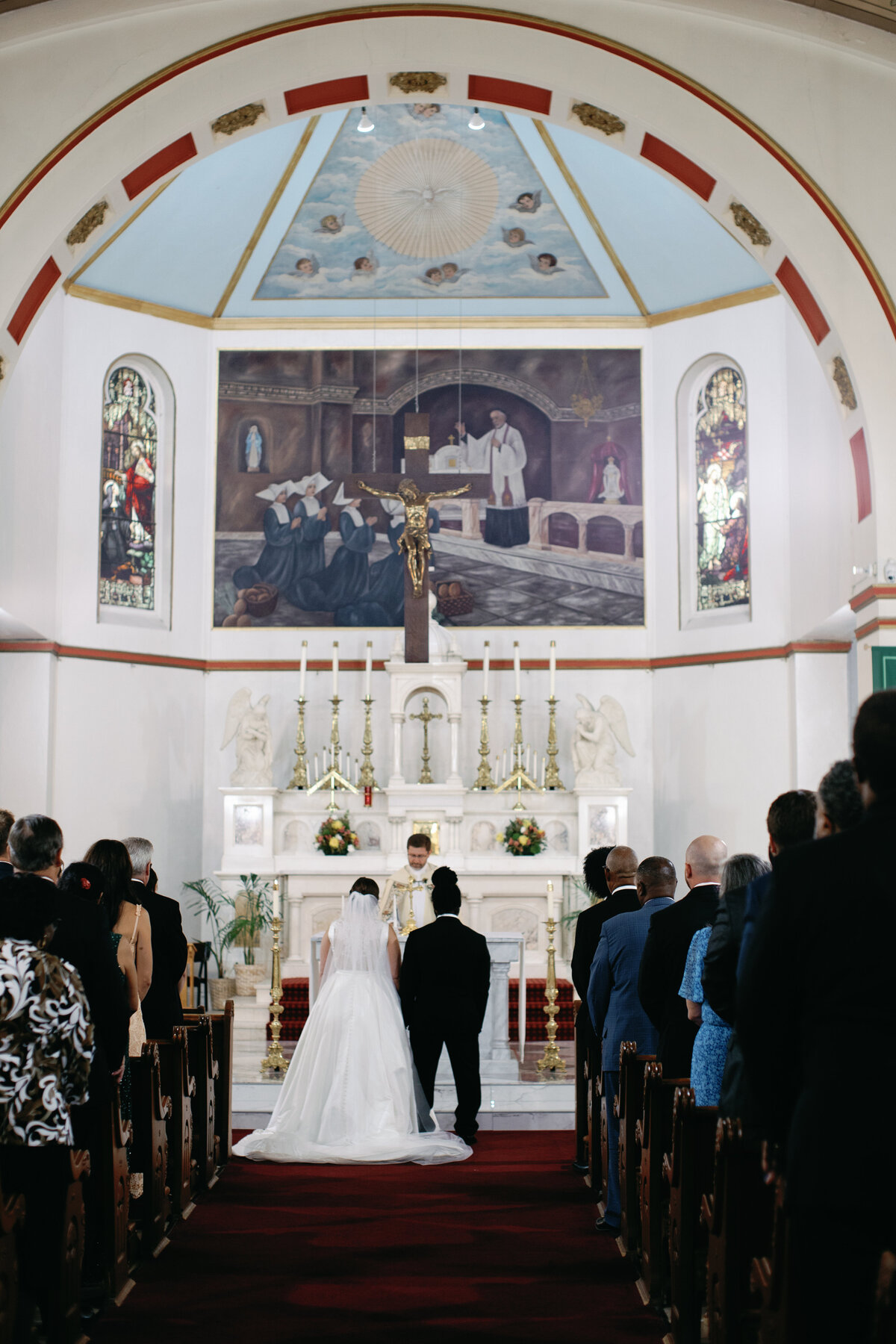 Bride and groom standing at the altar during Catholic wedding ceremony in Washington DC with ornate murals and stained glass.
