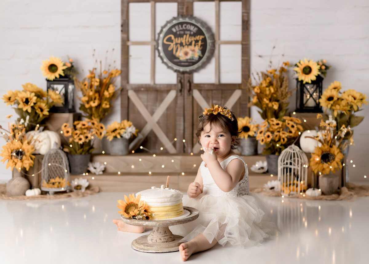 Rustic sunflower cake smash in West Palm Beach and Wellington photography studio. Baby girl in a white tulle dress is taking her first bite of cake. There is icing on her hands and face and she is looking at the camera. In the background are sunflowers and other yellow flowers against a rustic barn door.