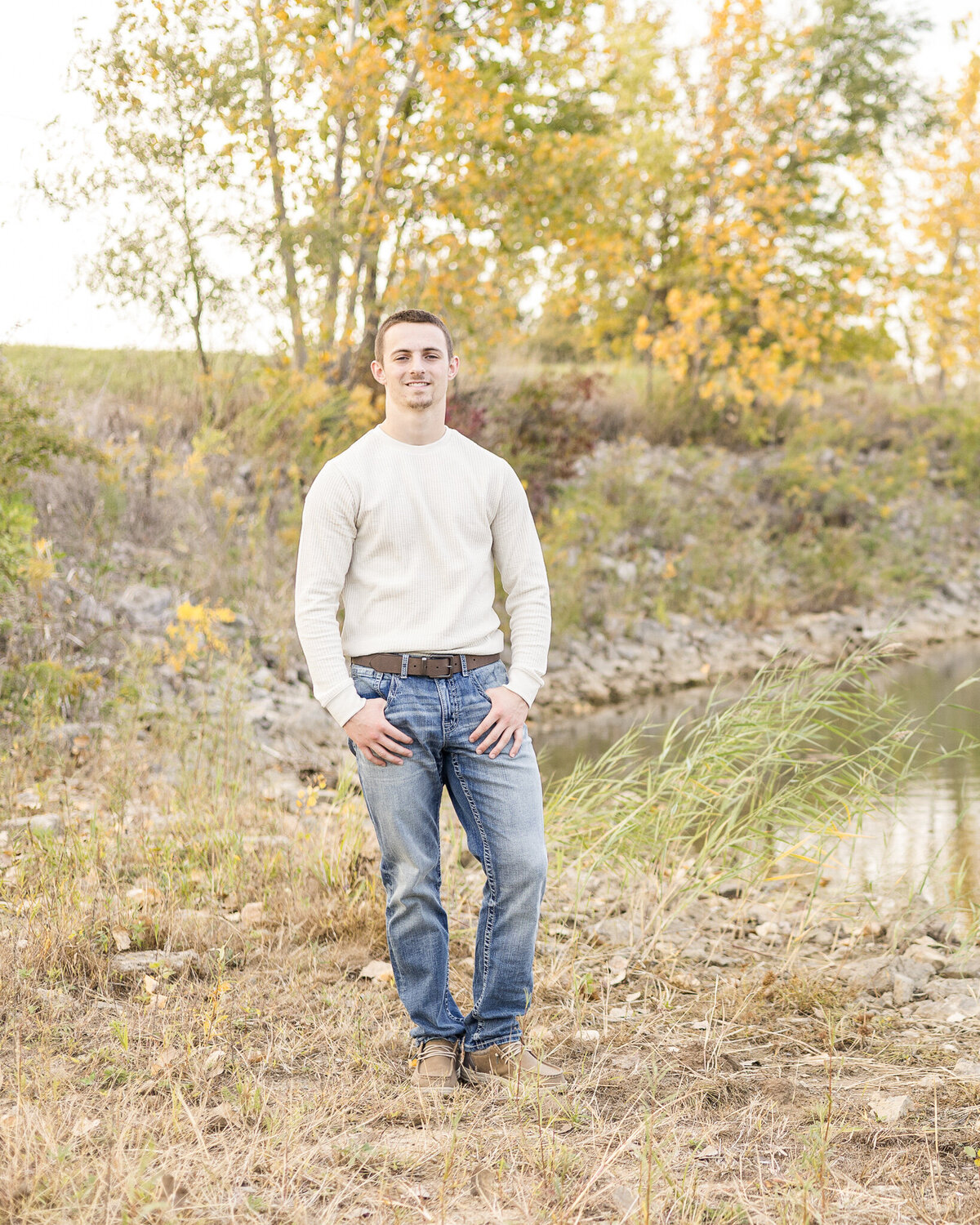High school senior male, standing next to the pond, with trees in the background, smiling at the camera, wearing a light tan long sleeve shirt, blue jeans, brown shoes with hands in his pockets.