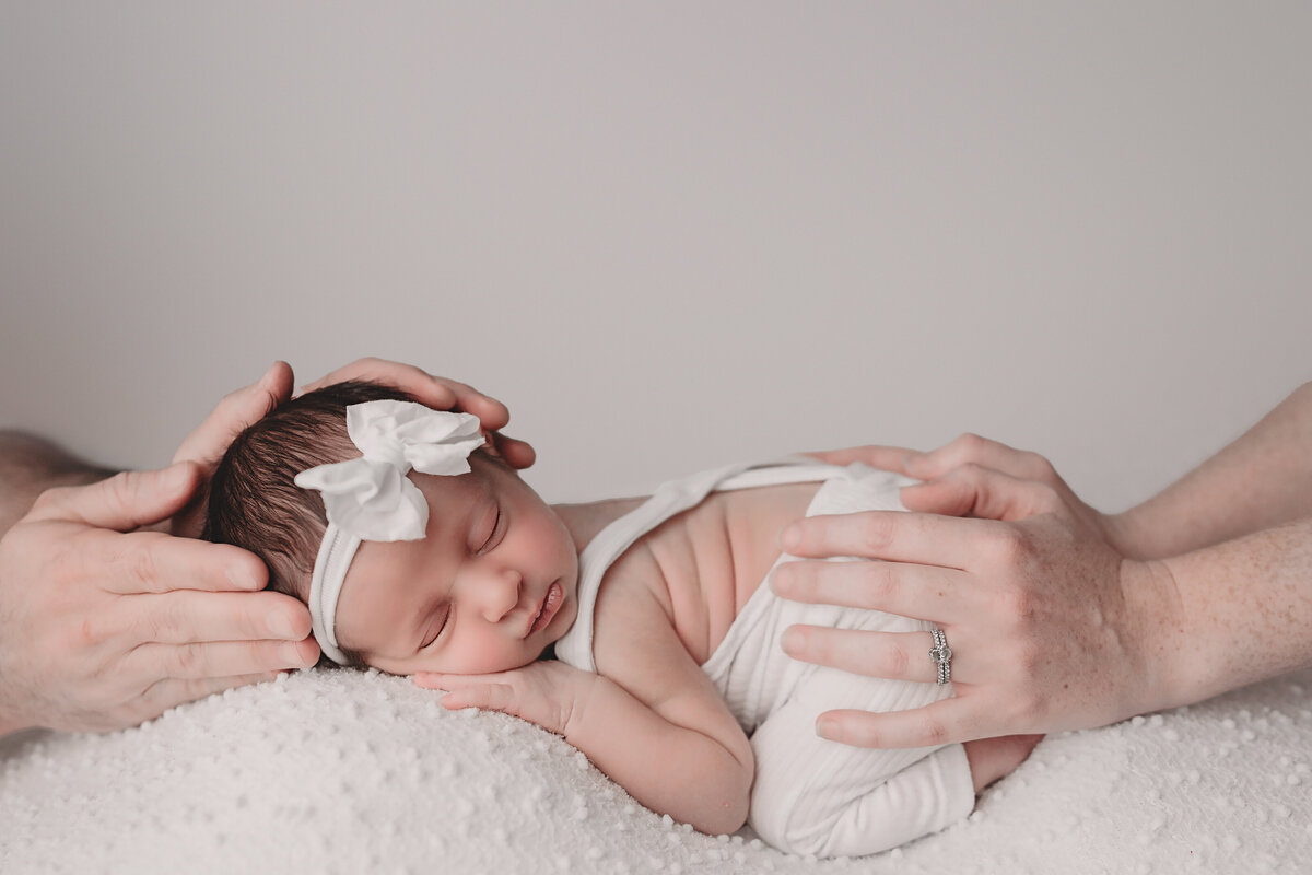 2 week old newborn girl posing on tummy down pose wearing white overalls and white bow with parents hands on her in front of white backdrop