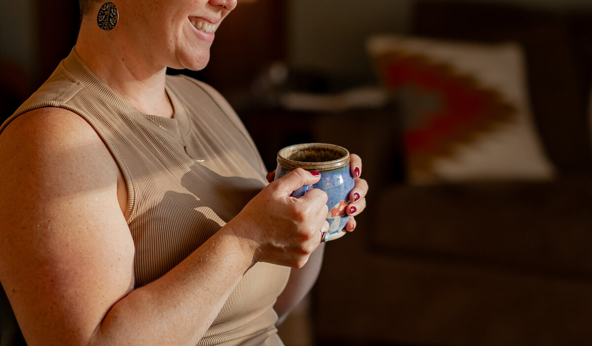 A person wearing a sleeveless top smiles while holding a blue mug, captured beautifully in this portrait photography. They are seated indoors with a blurred couch and pillow visible in the background, as sunlight casts soft shadows across the scene, evoking a cozy New England vibe.