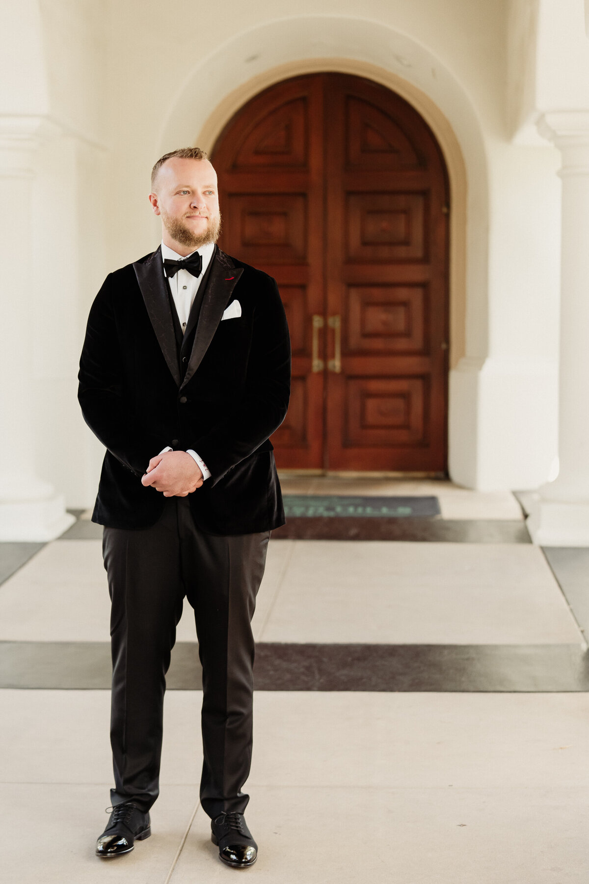 groom wearing a formal black velvet tux with a bowtie and leather and velvet shoes waits for his first look with his bride in front of large grand doors at spanish hills country club wedding captured by los angeles wedding photographer magnolia west photography