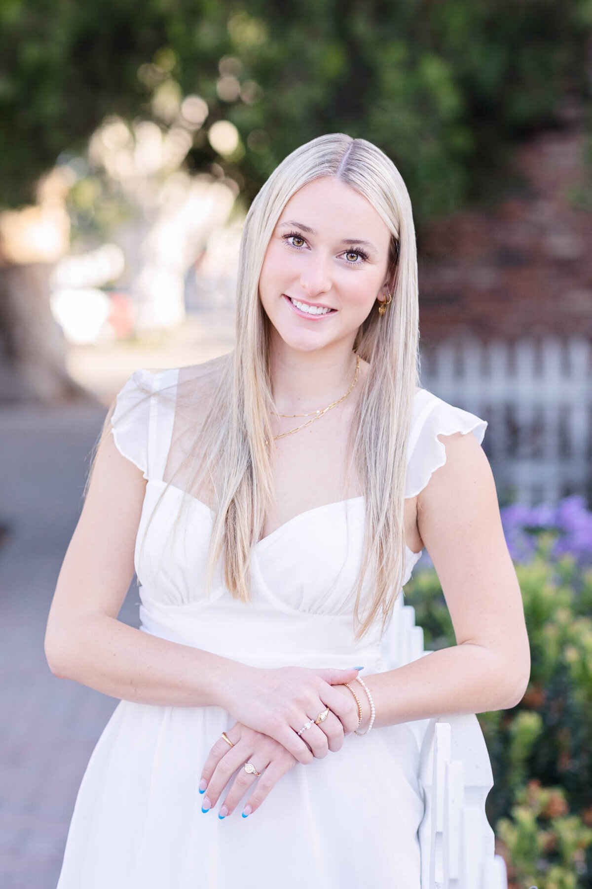 A woman with long blonde hair stands on a street, leaning on a white fence. She is wearing a white dress and smiling. There are trees and a brick wall in the background.