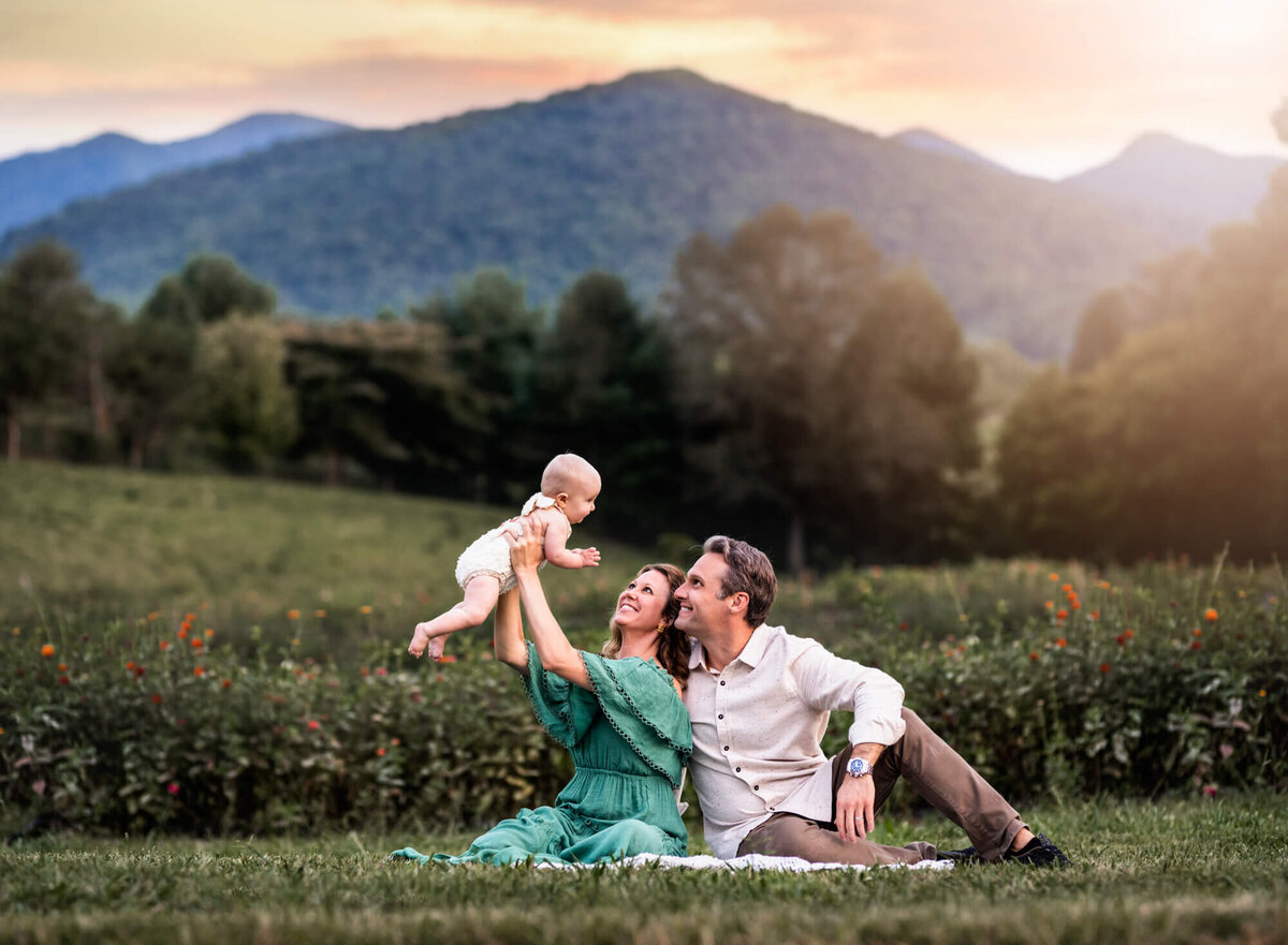 An adorable mom and dad hold up their baby girl in front of a filed of flowers with mountains in the distance