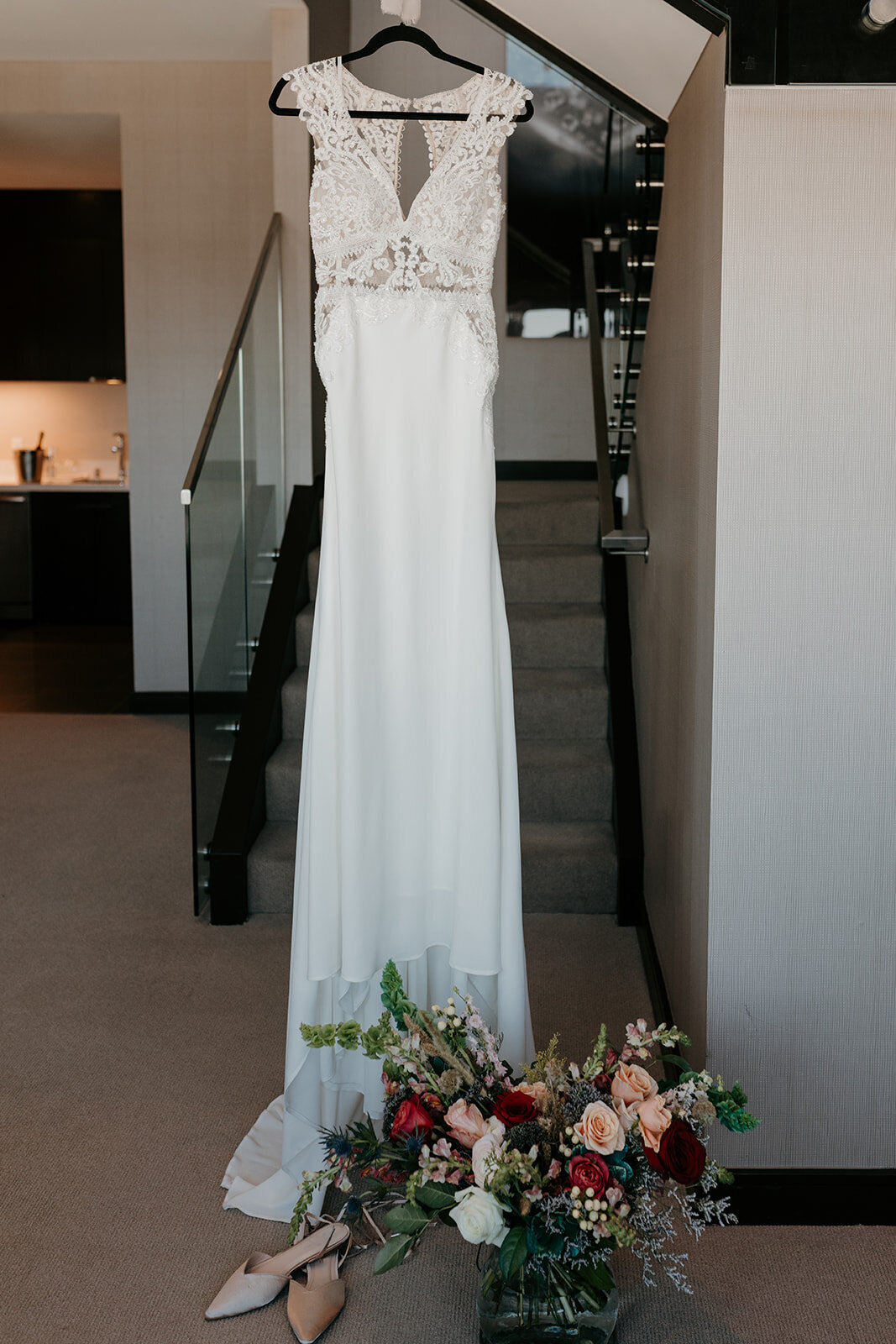 A sleeveless lace wedding gown on a hanger, displayed against a staircase, with a vibrant bouquet of mixed flowers and pointed-toe shoes placed on the floor below, capturing a moment before the ceremony.