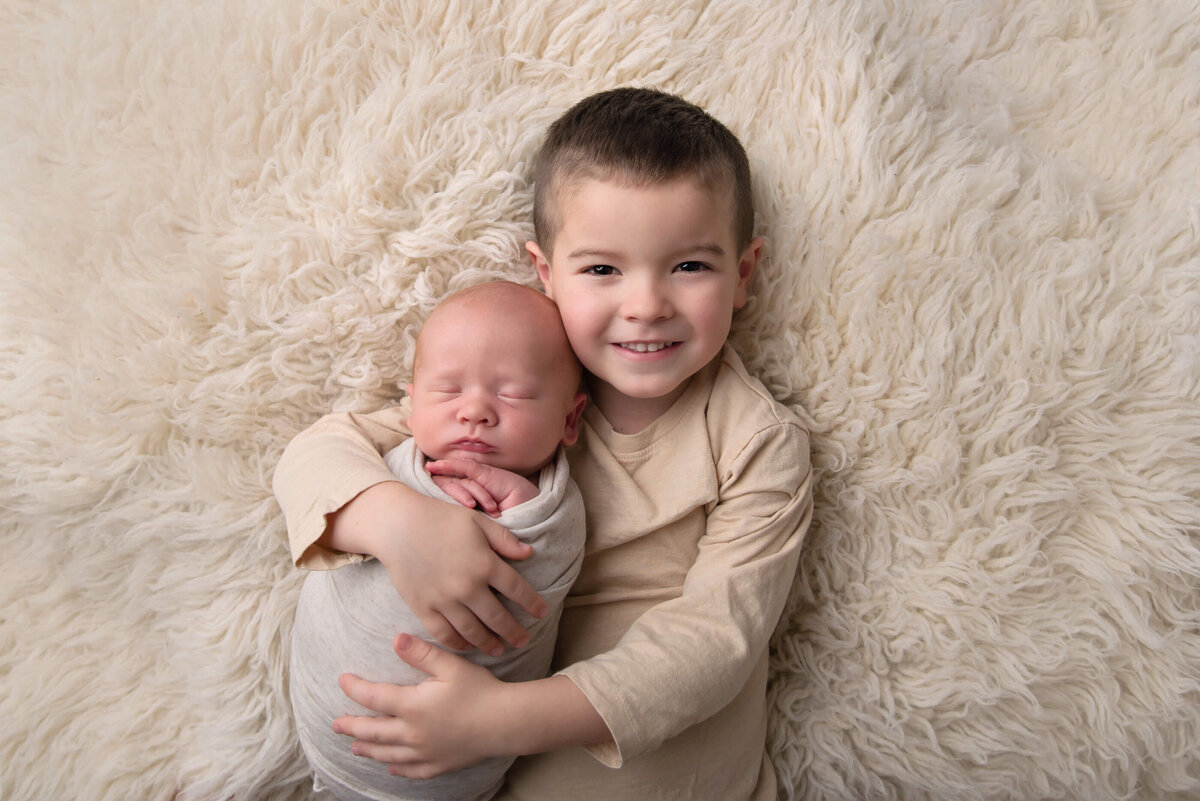 big brother holding newborn baby brother laying on a cream flokati for their newborn photography session in Burlington, Ontario