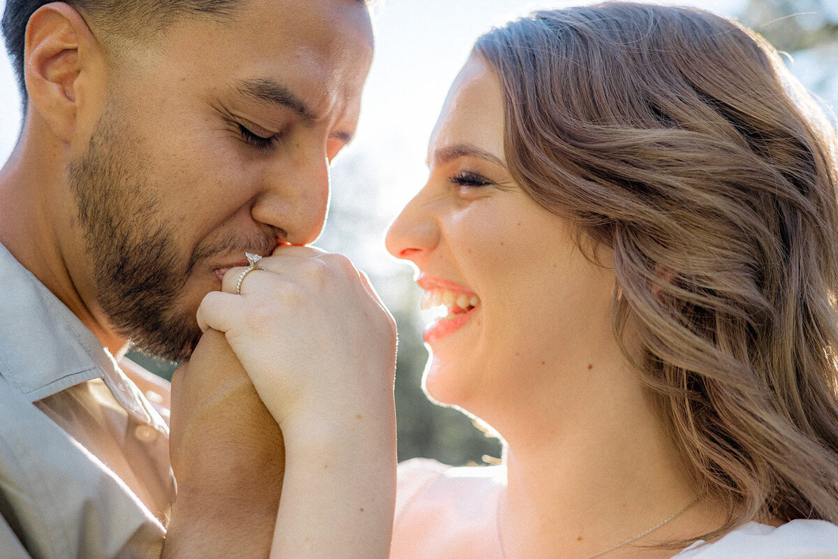 Close-up of the couple, where the man kisses the woman's hand as she smiles, displaying her engagement ring.