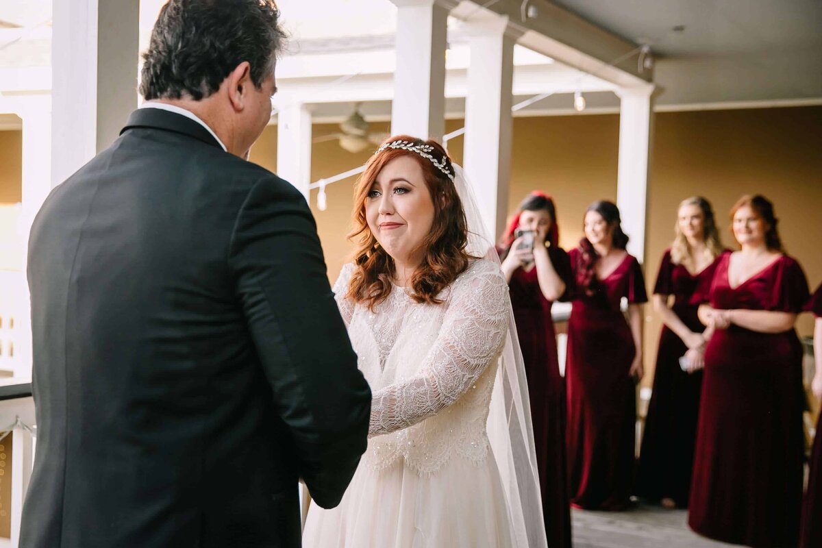 A bride in a white dress, appearing emotional, holds hands with her father during a first look. Bridesmaids in burgundy dresses watch and smile in the background on a porch.